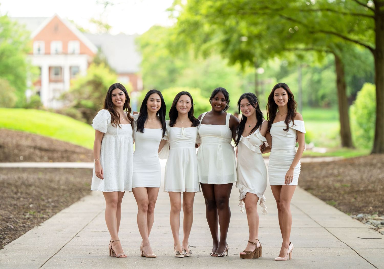 Group of graduates in white dresses posing together on campus walkway.