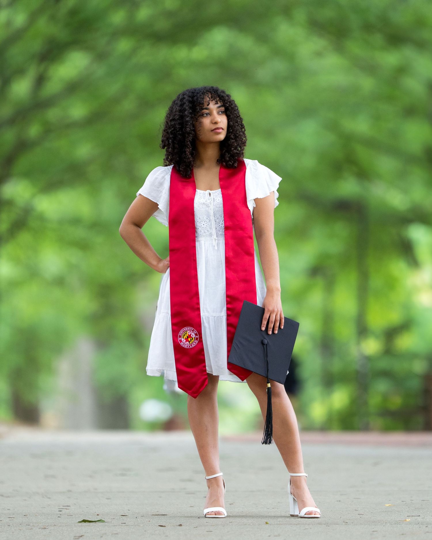 Graduate in white dress and red stole holding diploma case on tree-lined path.
