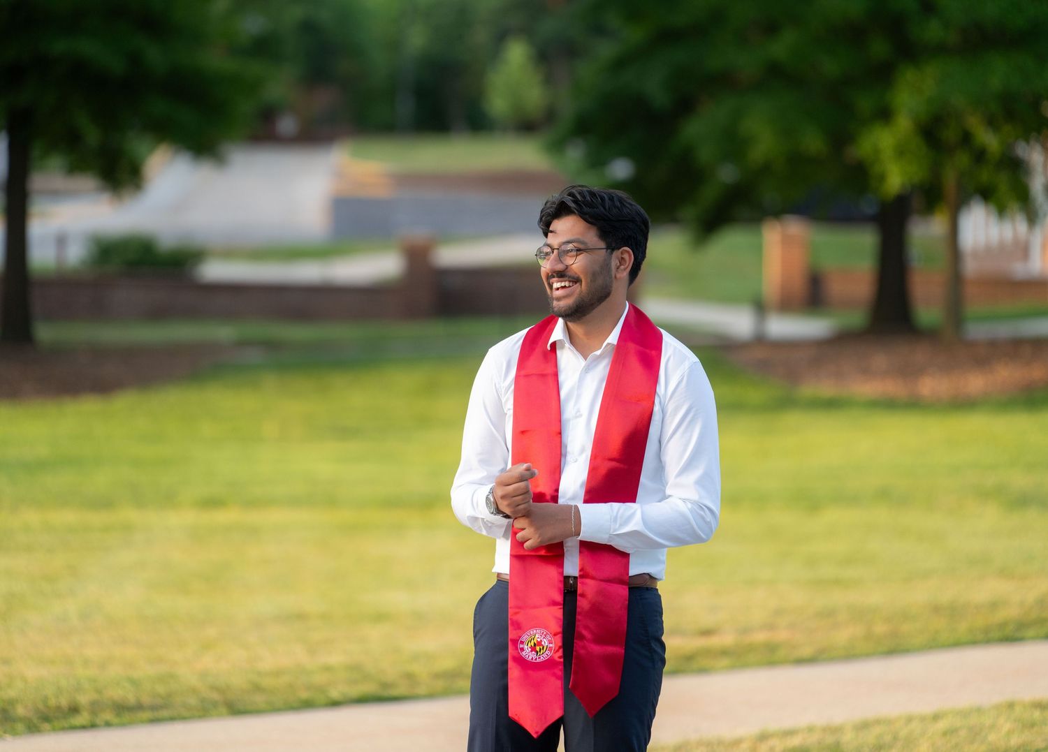 Graduate in formal attire with red stole standing on campus lawn.