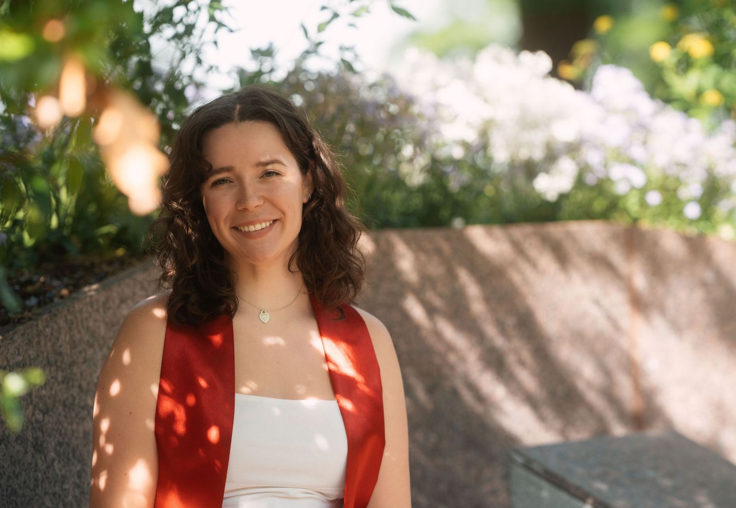 Graduate in red and white regalia smiling in dappled sunlight.