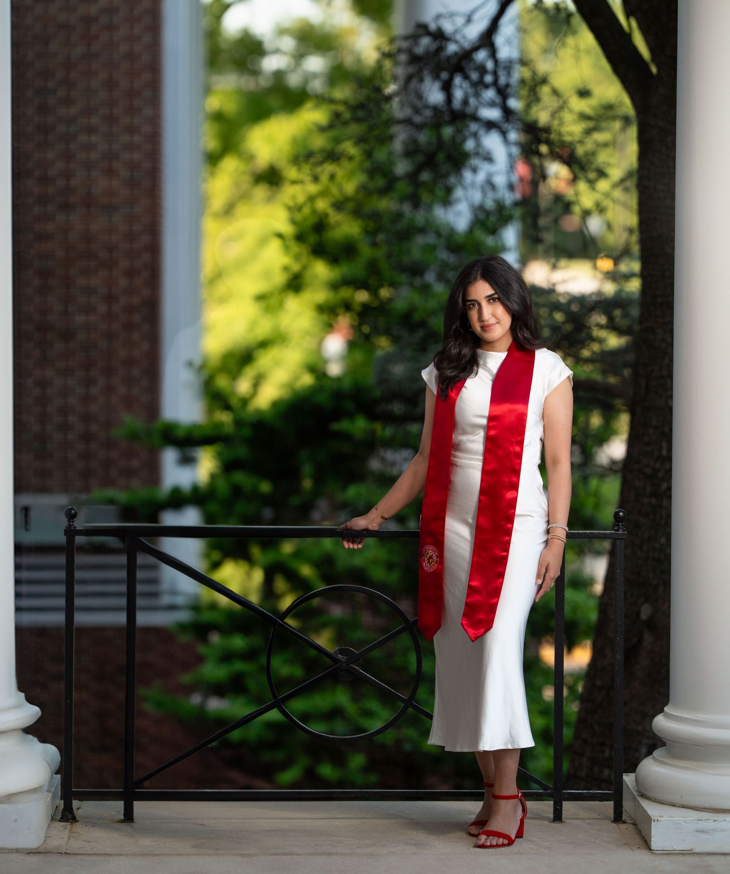 Graduate in white dress and red stole standing near classical columns.