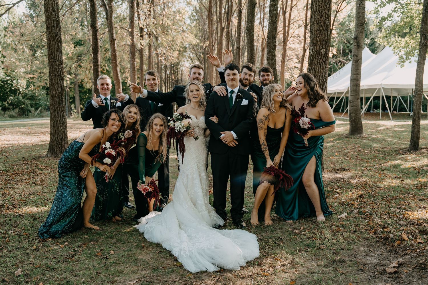 Wedding party in teal dresses and black suits pose together in a wooded outdoor setting with a white tent in background.
