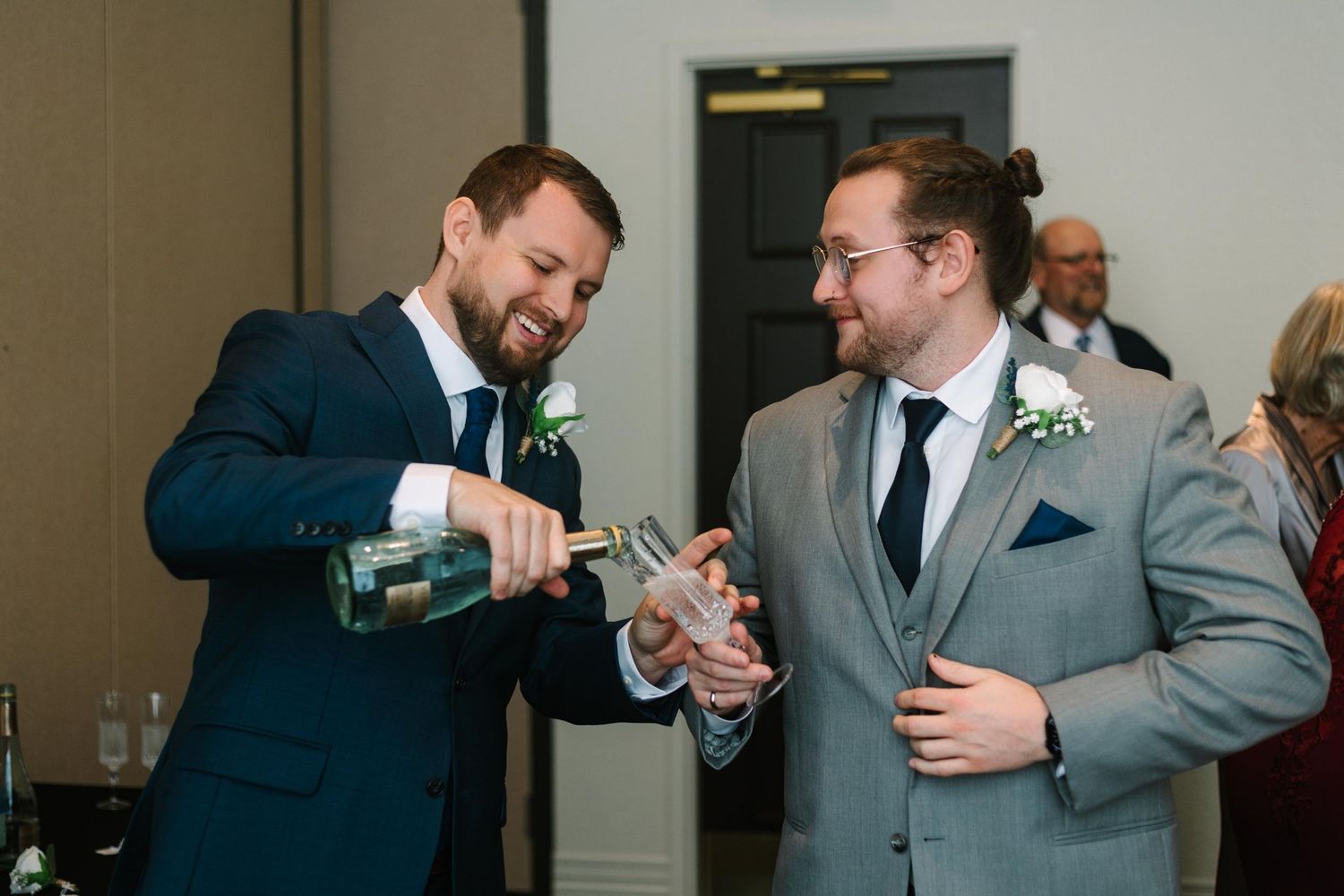 Two men in navy suits share a laugh while pouring drinks at a formal event.