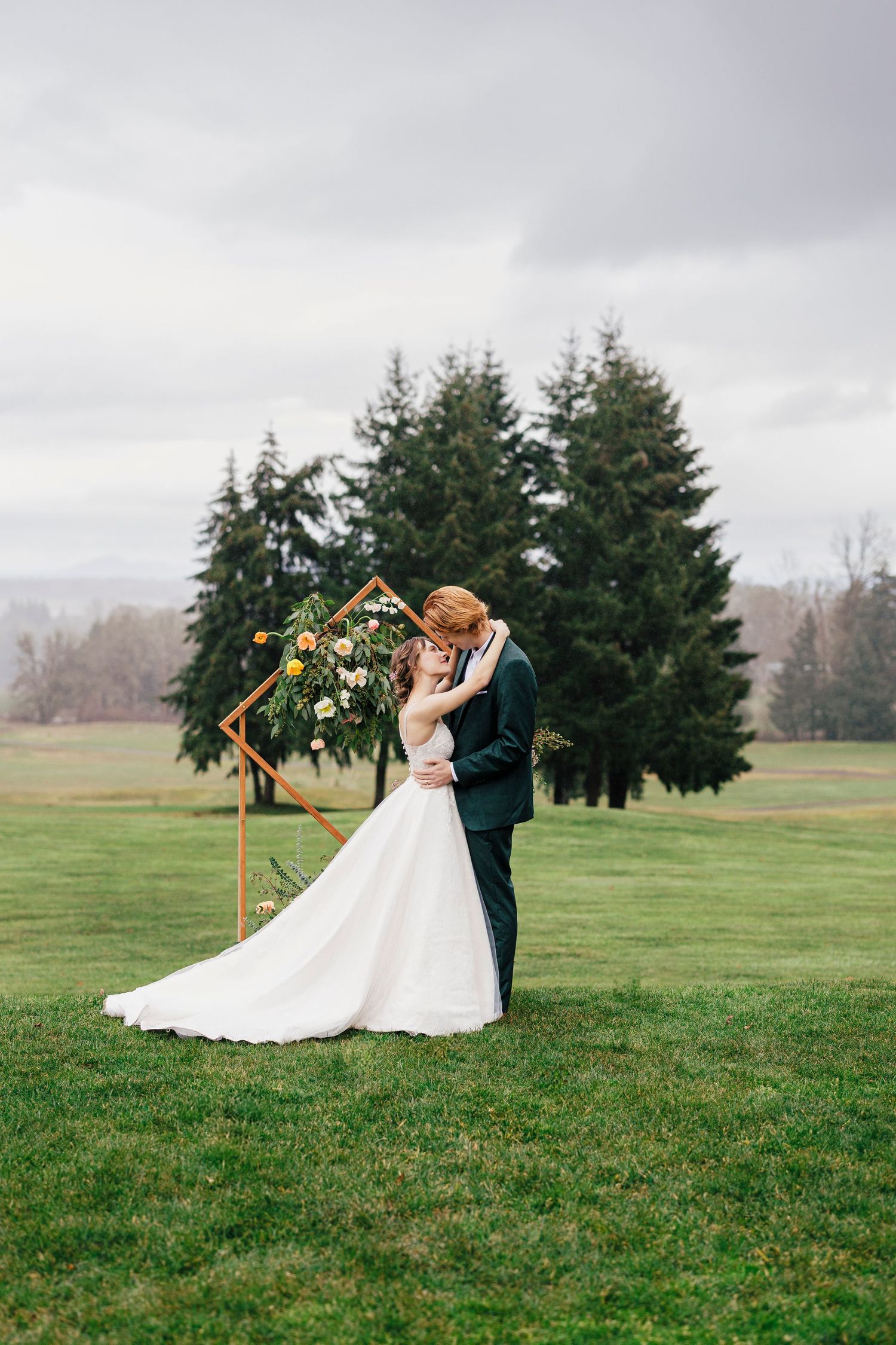 A couple shares an intimate moment during their wedding ceremony under a wooden arch on a lush green field.