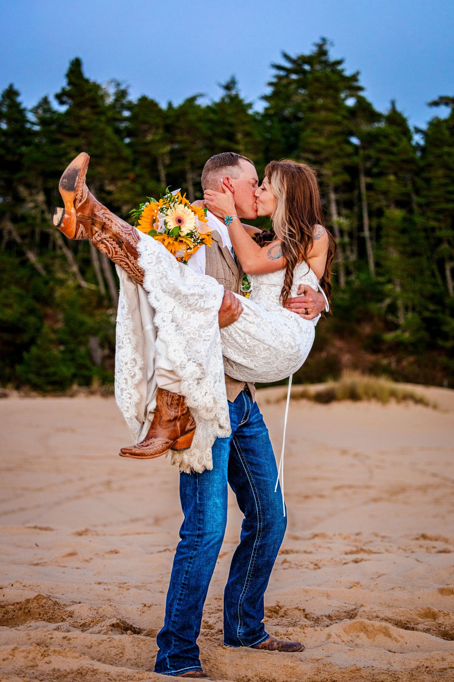Groom lifts bride in white dress and cowboy boots for romantic beach kiss.