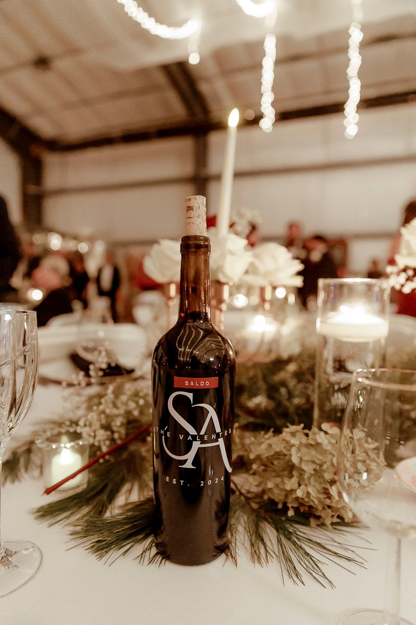 A wine bottle on an elegant wedding reception table decorated with pine branches and twinkling lights.