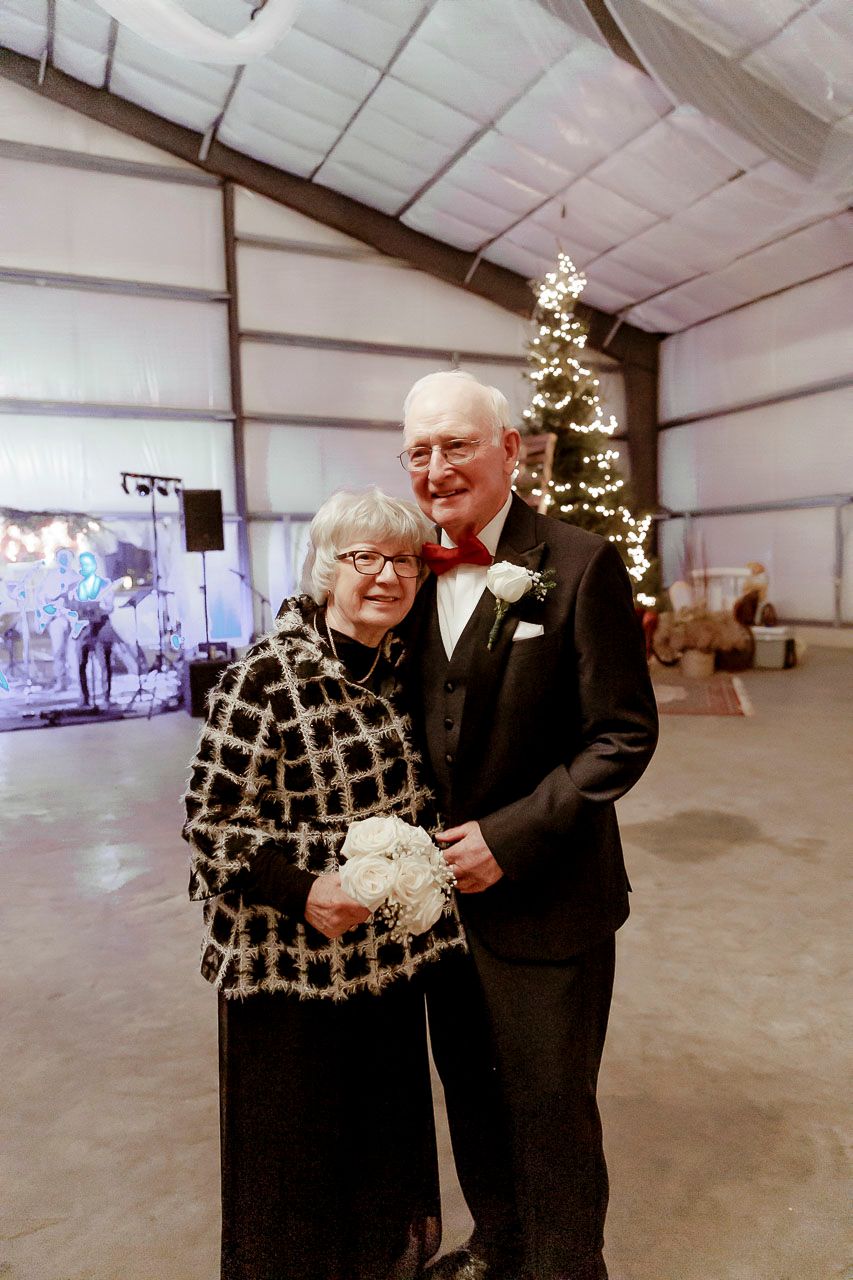 Elderly couple smiles together at holiday celebration in decorated barn-style venue with lit Christmas tree in background.