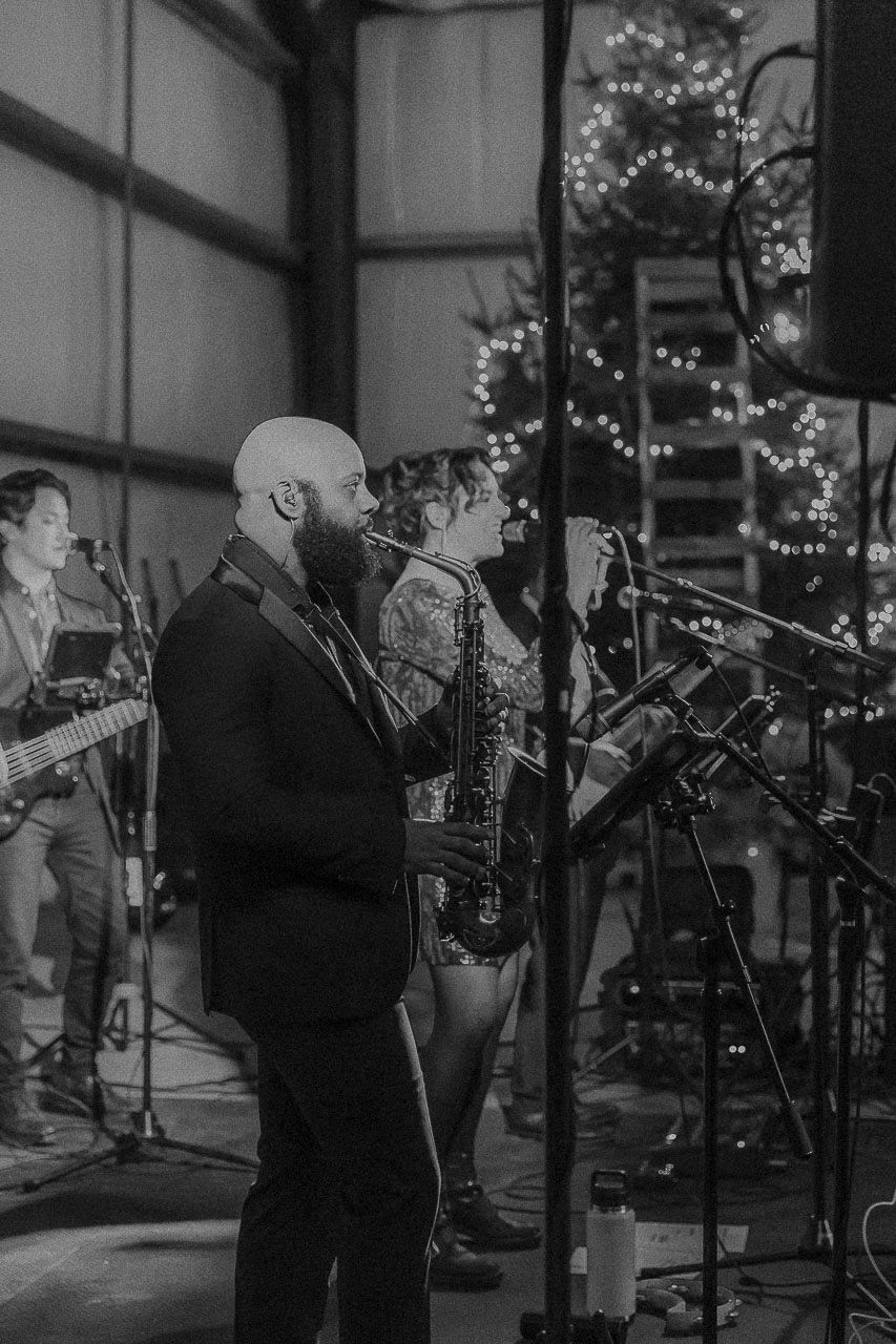 Black and white photo of a bearded performer singing on stage with musicians playing instruments and holiday decor in background.