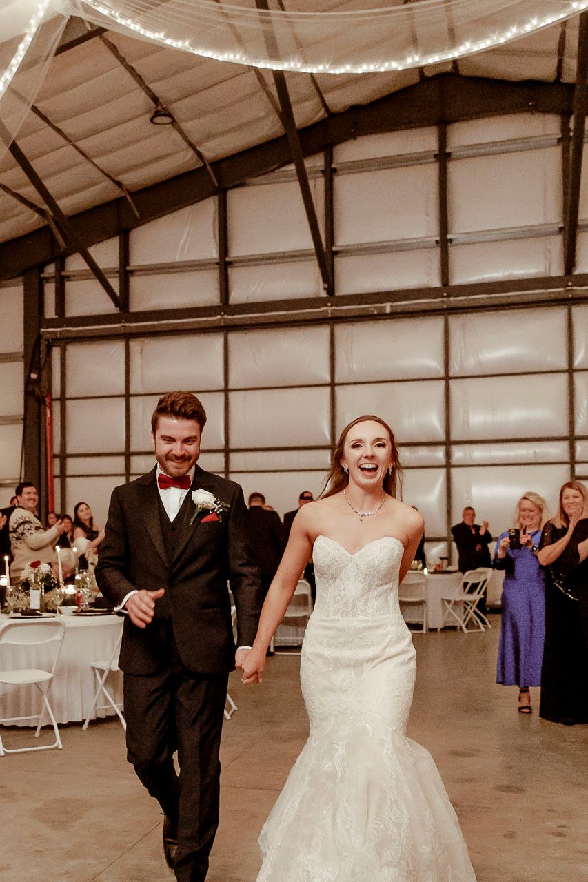 Joyful couple makes their grand entrance at a wedding reception in an industrial venue decorated with string lights.