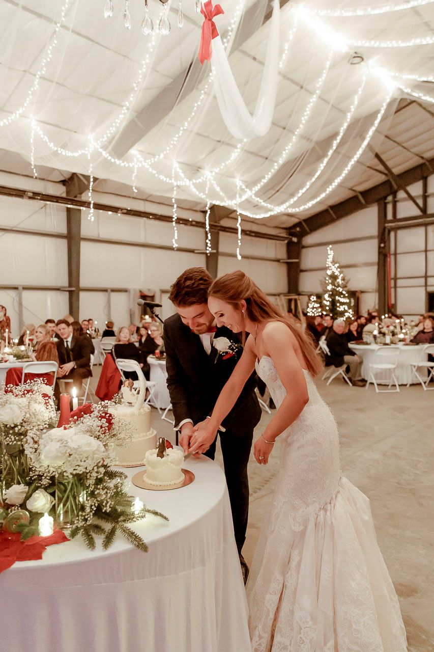 A couple cuts their wedding cake together at a rustic reception venue decorated with white draping and twinkling lights.