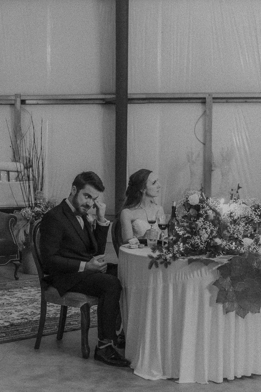 A black and white wedding photo showing couple seated at decorated table with floral arrangements in an indoor venue.