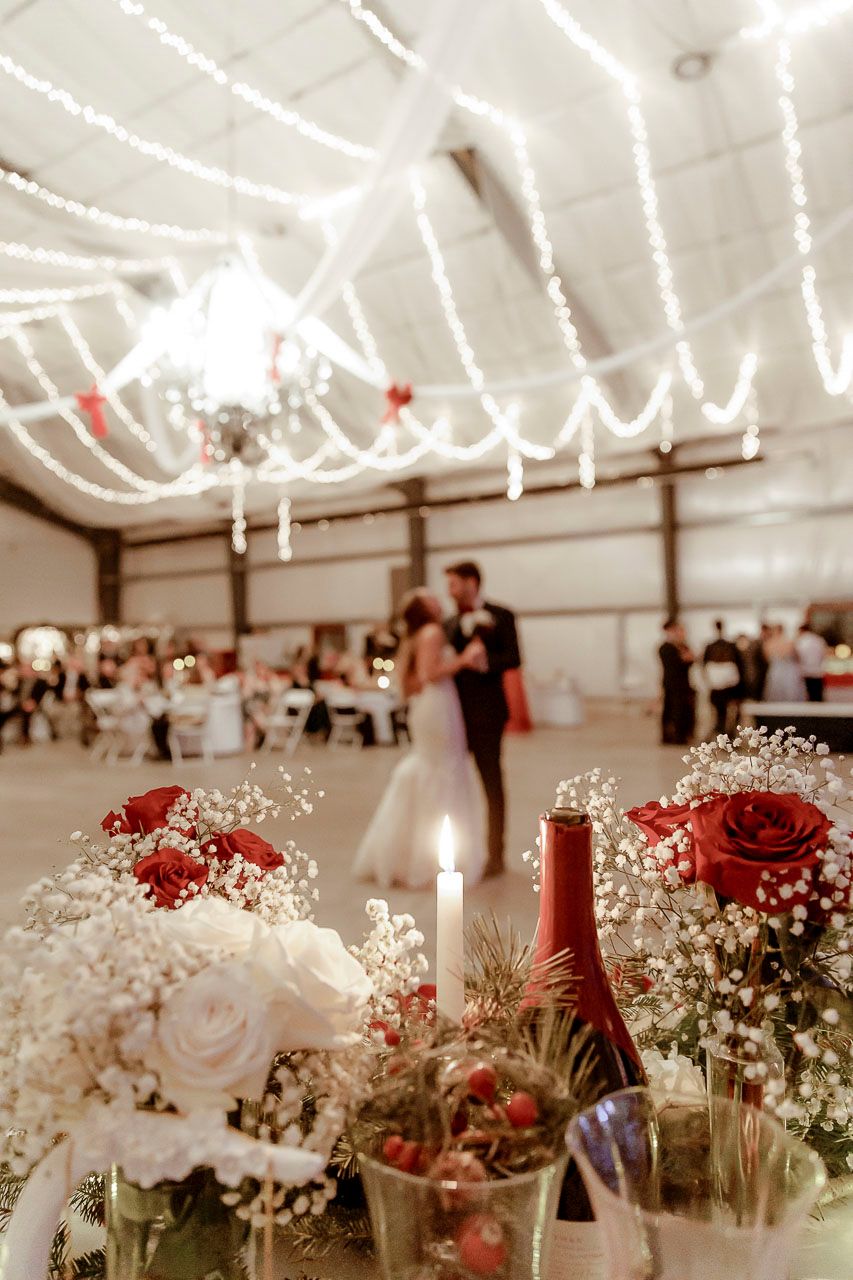 Reception decorations of red and white roses with baby's breath and wine bottles under twinkling string lights in an indoor venue.
