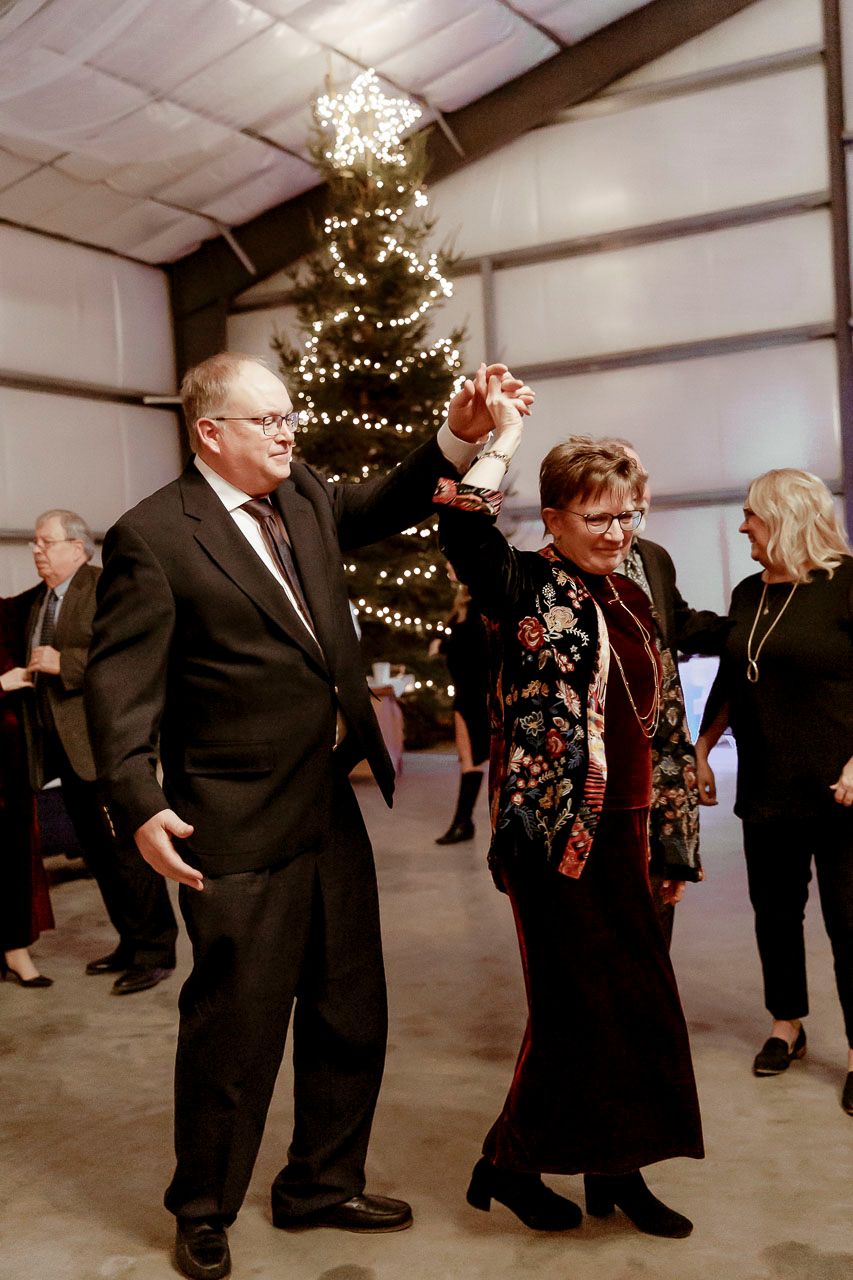 Guests dance during a festive celebration with a lit Christmas tree and other party-goers in the background.