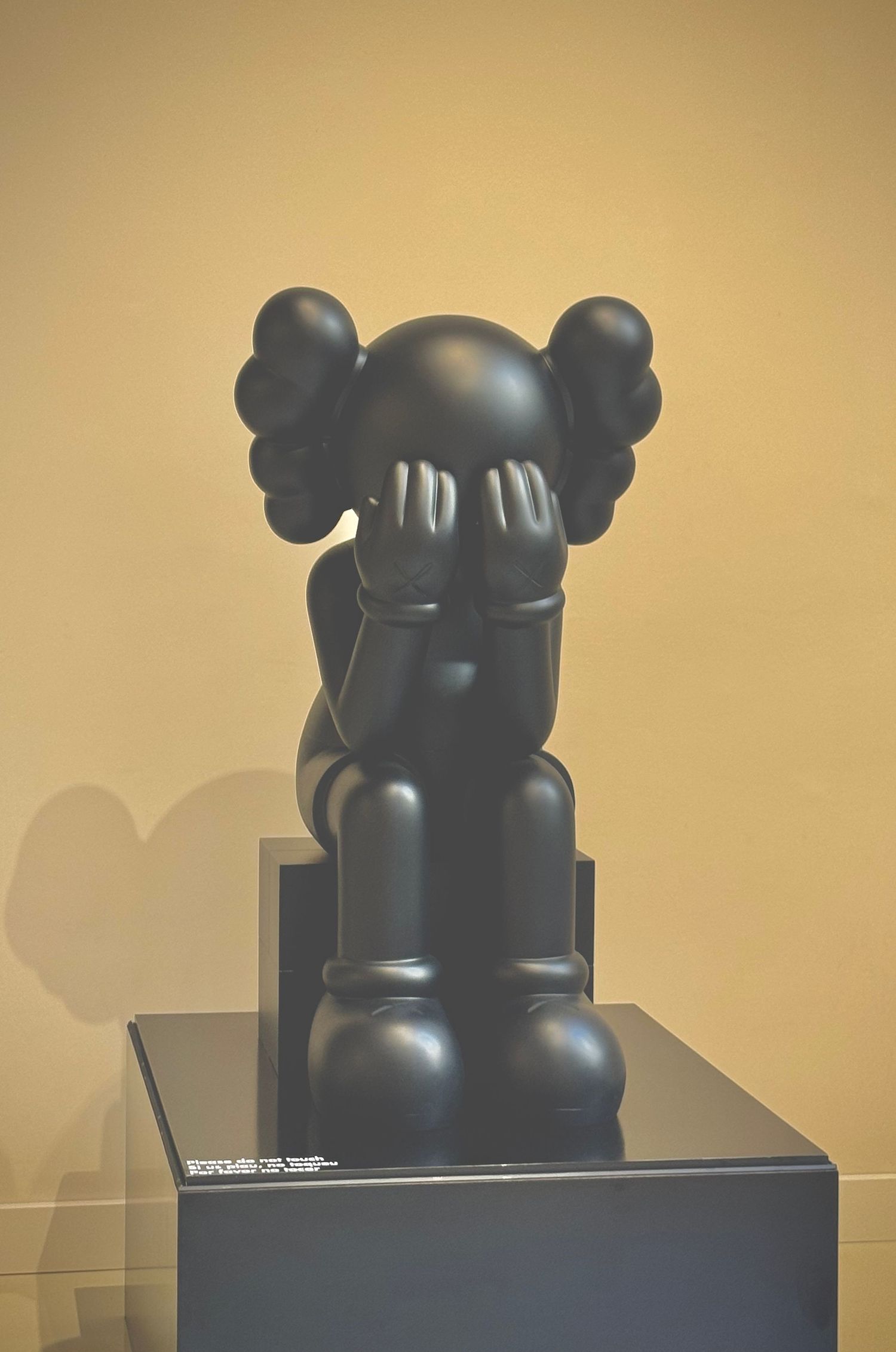 Black KAWS figurine sculpture displayed on pedestal against beige background.