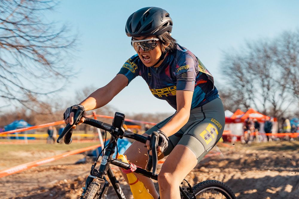 Cyclist in blue jersey races on dirt track during cyclocross competition with bare trees in background.