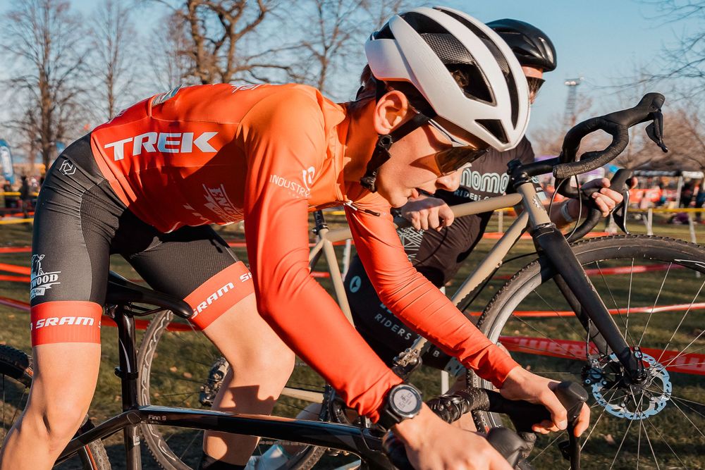 Cyclist in orange Trek team jersey races on a competitive cyclocross course during a sunny winter day.