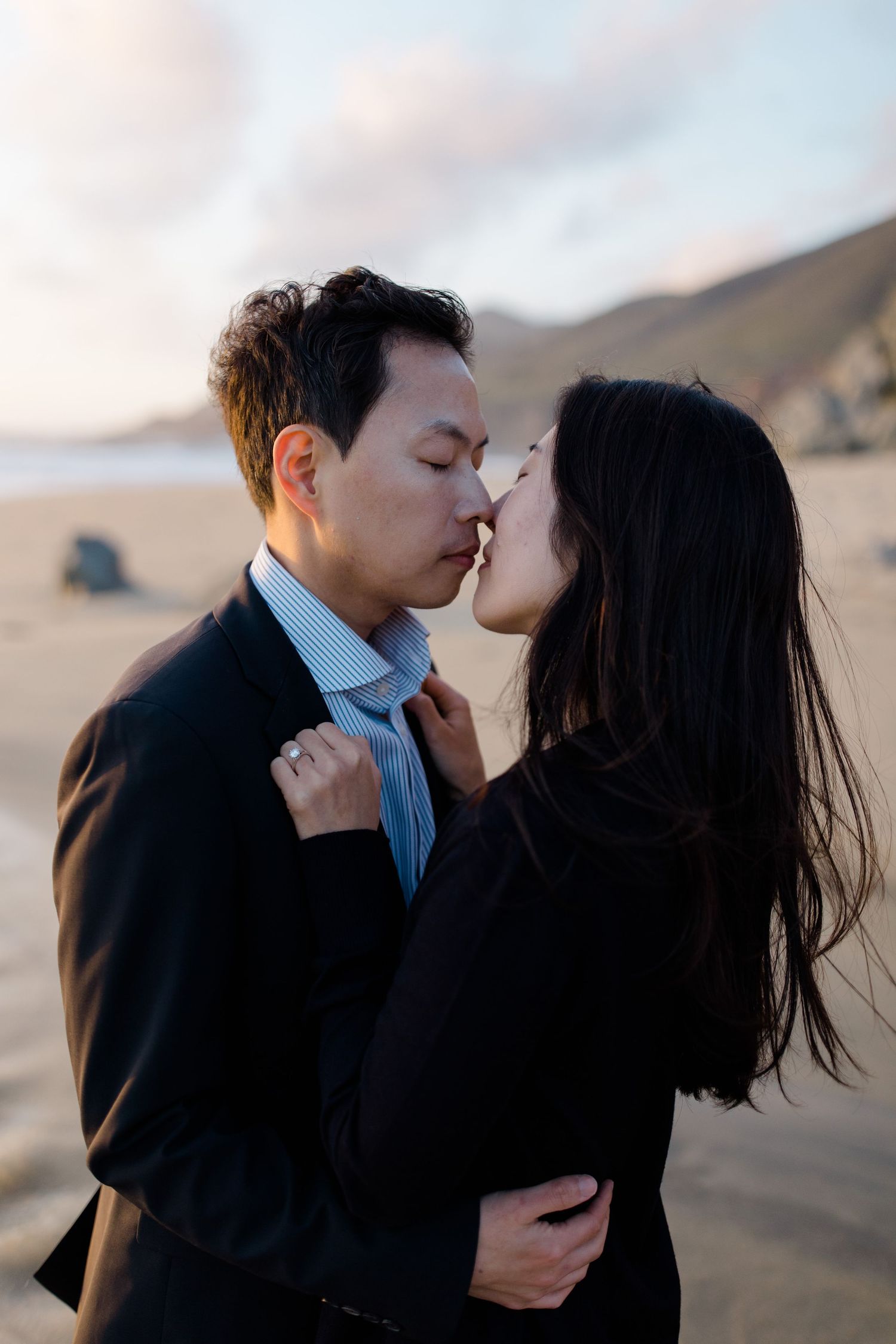 Romantic couple sharing an intimate kiss on a beach at sunset while dressed in formal attire.