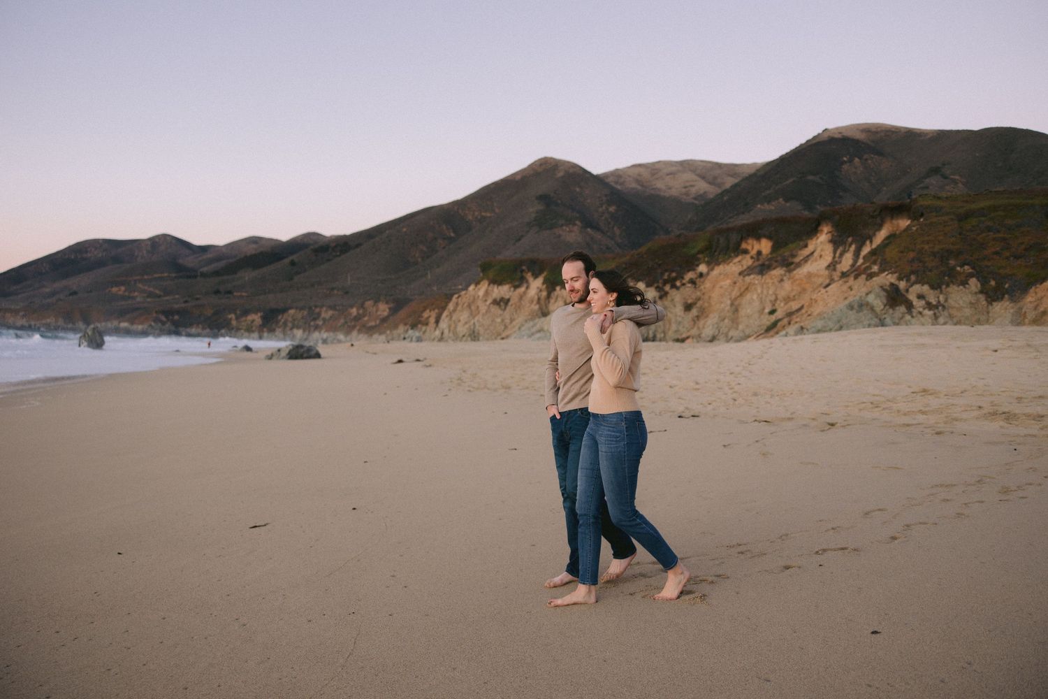 Two people embrace while walking along a scenic beach at sunset with mountains and cliffs in the background.