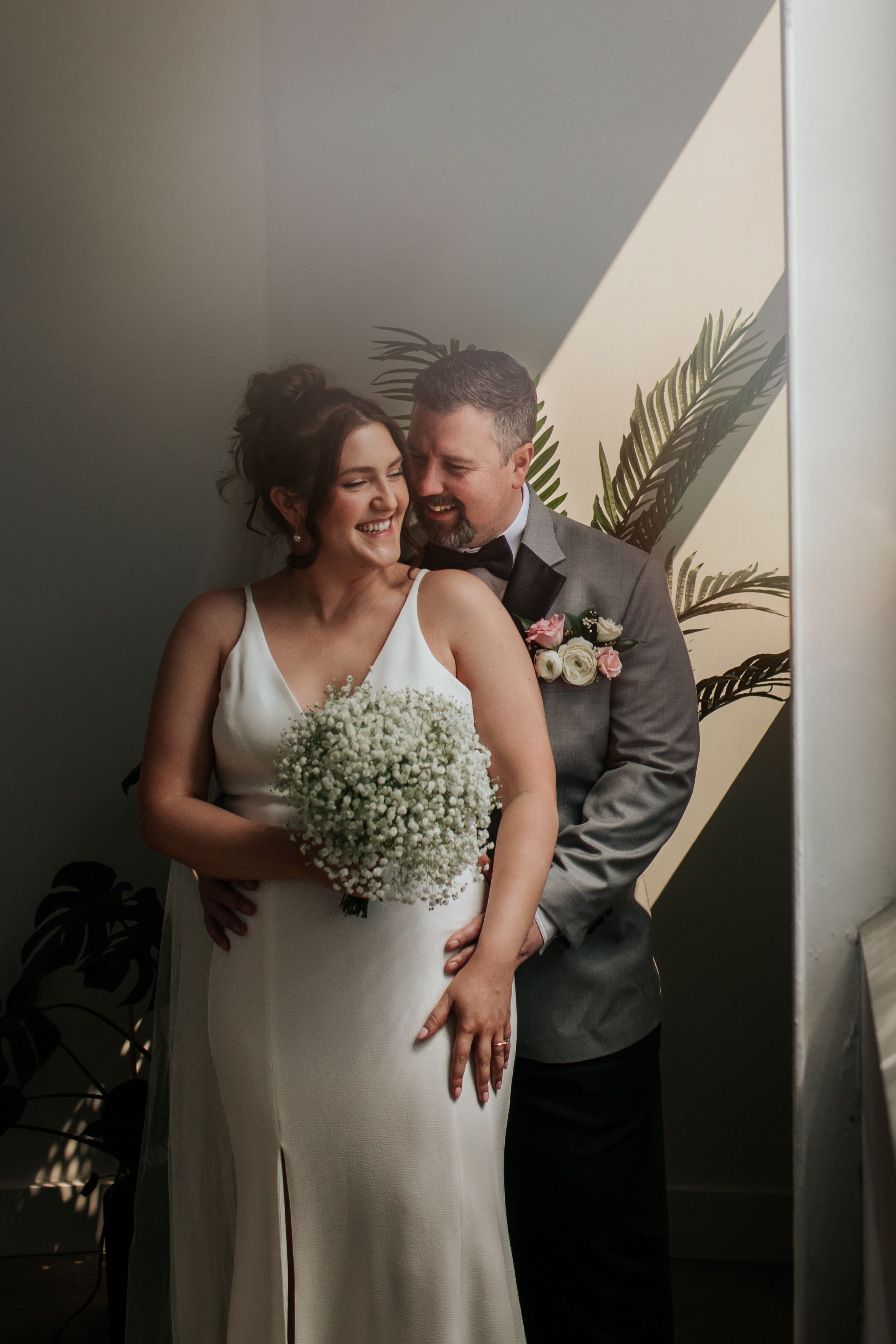Romantic wedding couple embraces in soft window light while holding delicate baby's breath bouquet in Indianapolis Indiana.