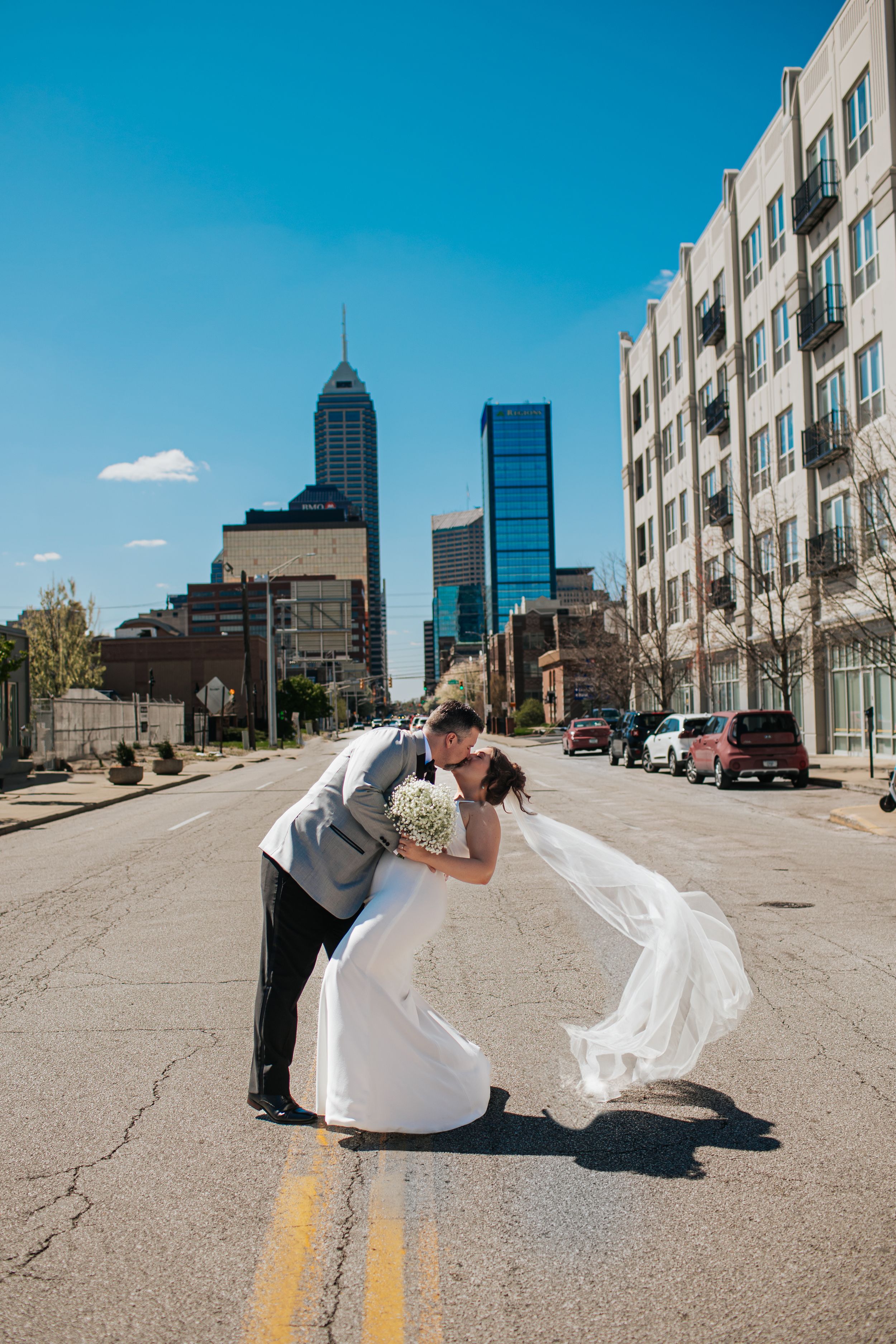 A romantic wedding couple dips and kisses on an urban street with the Indianapolis skyline in the background in Indianapolis Indiana.