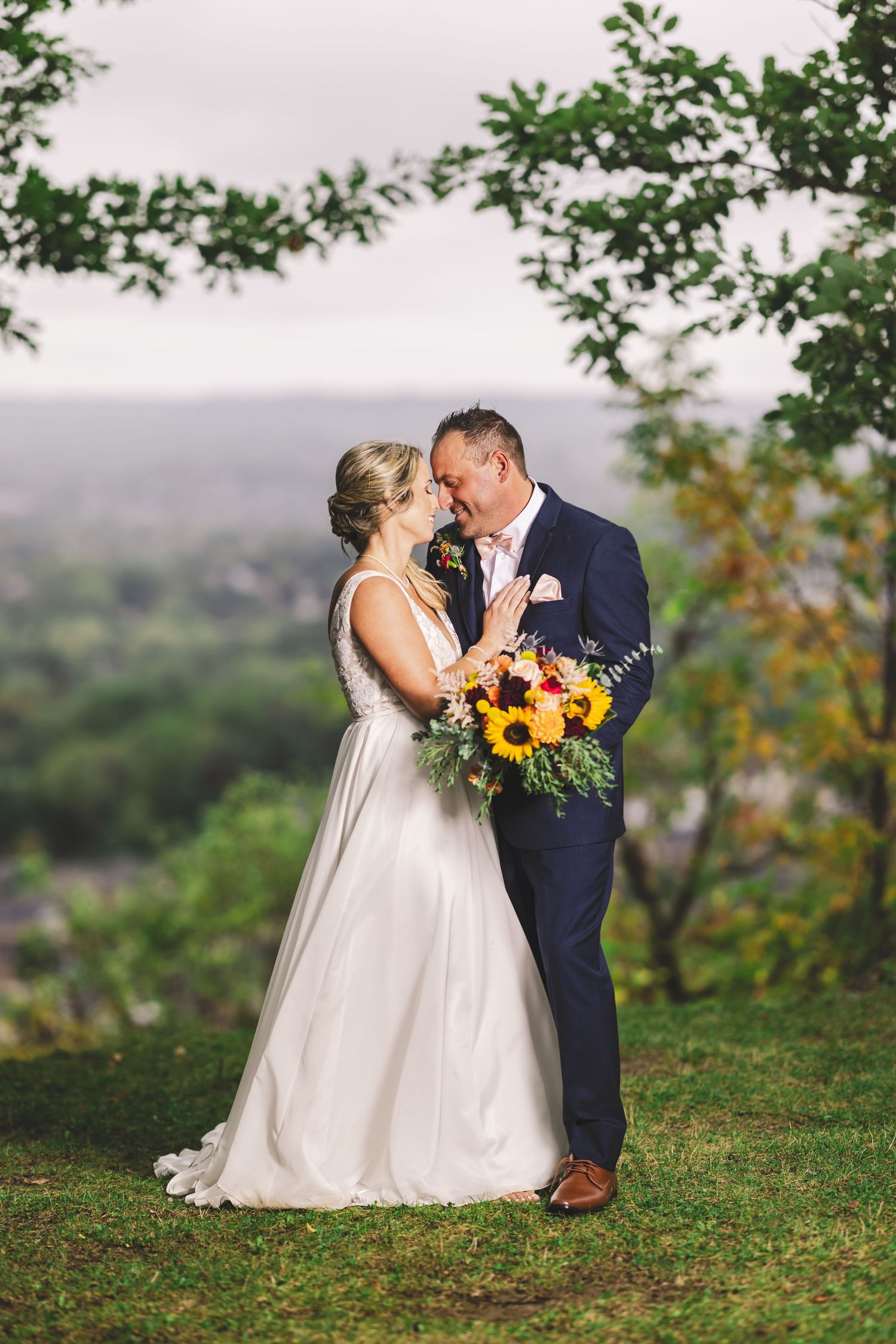 Couple in wedding attire embrace on hilltop with bright sunflowers and greenery in misty outdoor setting.