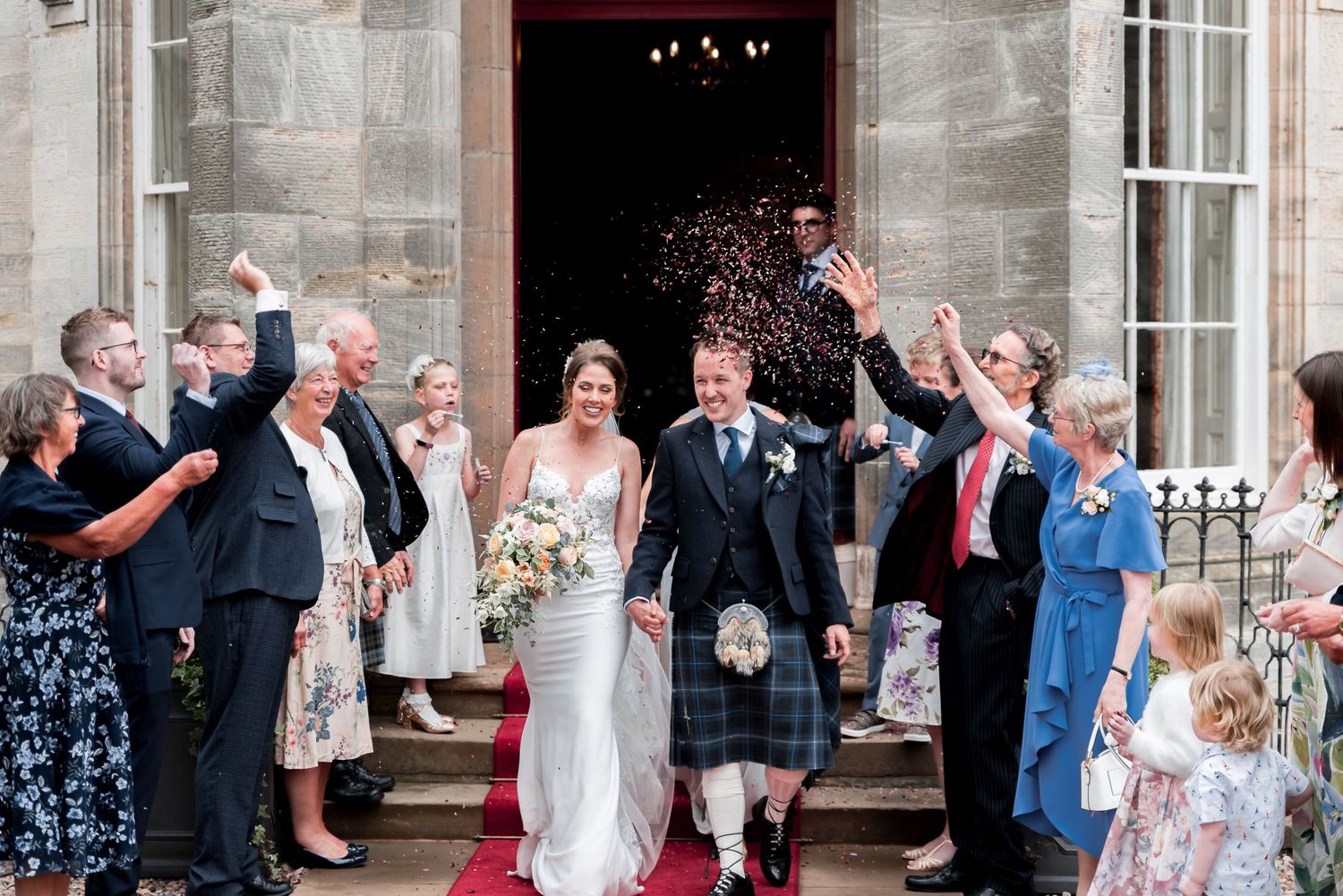 Wedding guests shower confetti on couple exiting stone church building in traditional Scottish attire.