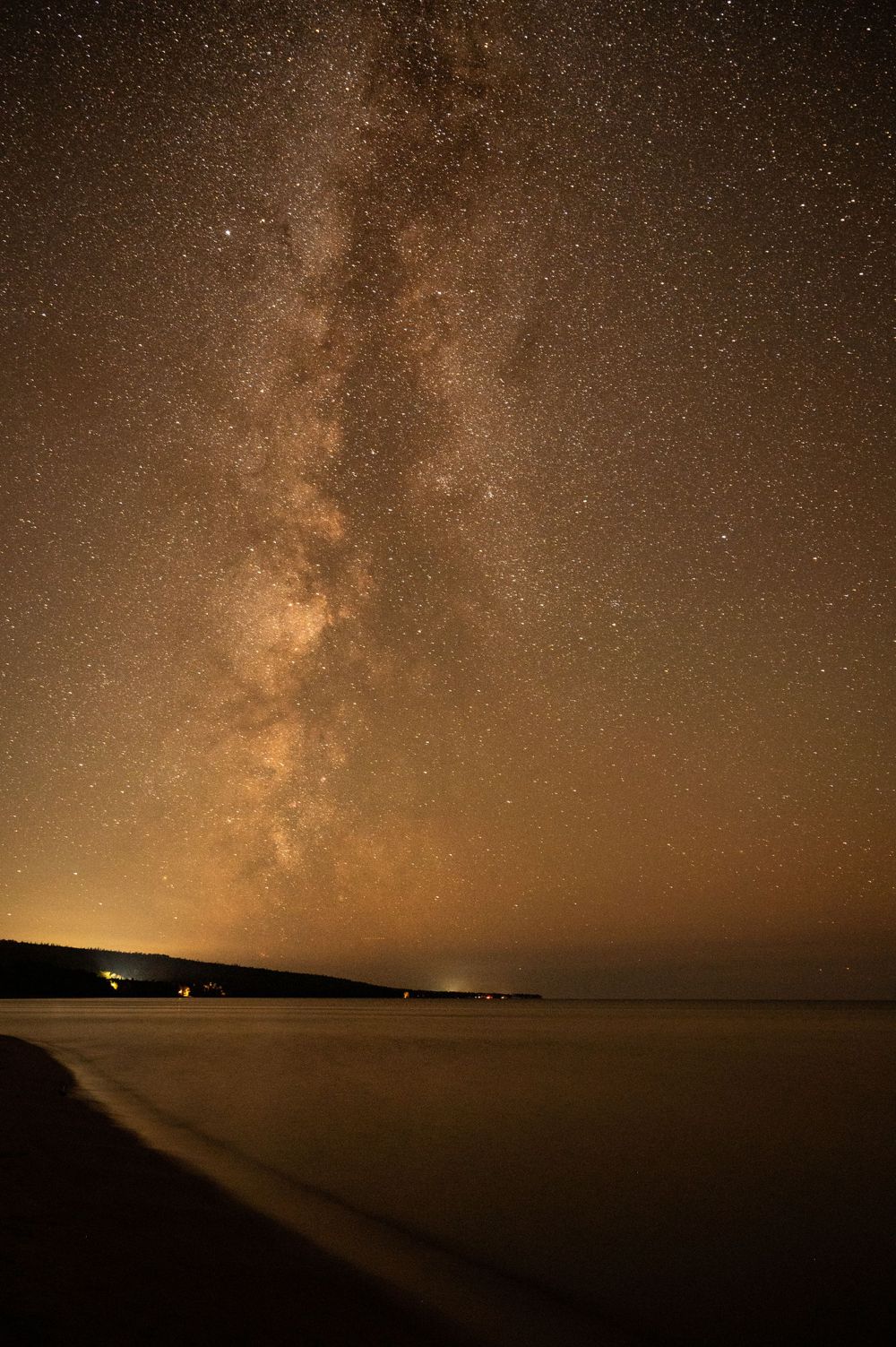 Stunning Milky Way galaxy illuminates the night sky over a tranquil shoreline with distant lights along the coast.