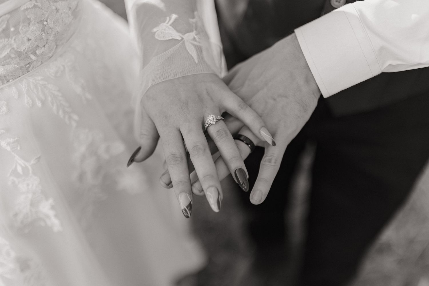 Close-up of wedding rings and hands against white lace dress fabric in black and white.
