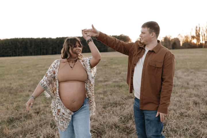 A couple smiles and dances together in a golden field at sunset, celebrating a special moment together before their new baby gets here.