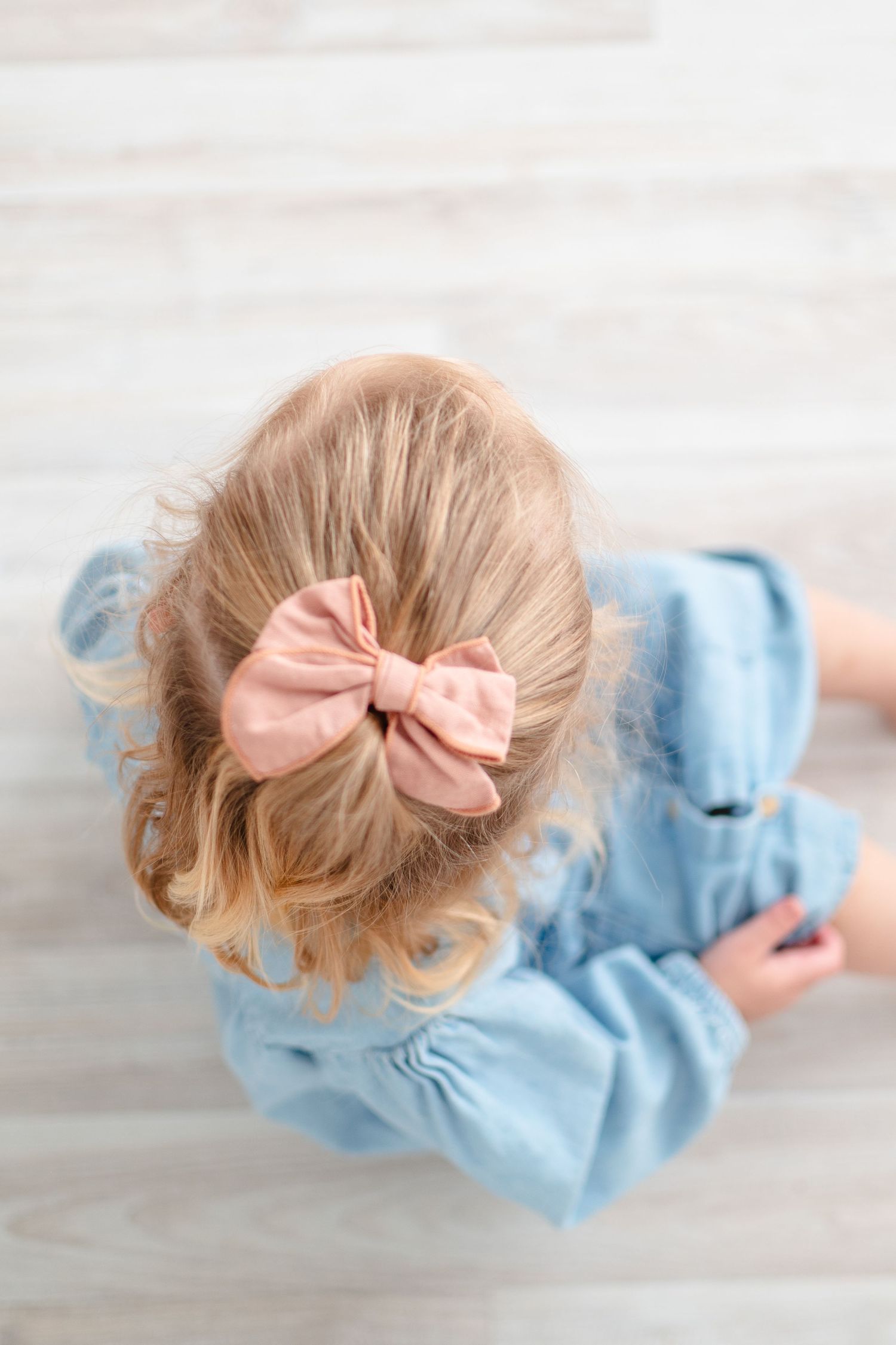 Overhead view of blonde hair styled with a pink bow and wearing light blue dress on white floor.