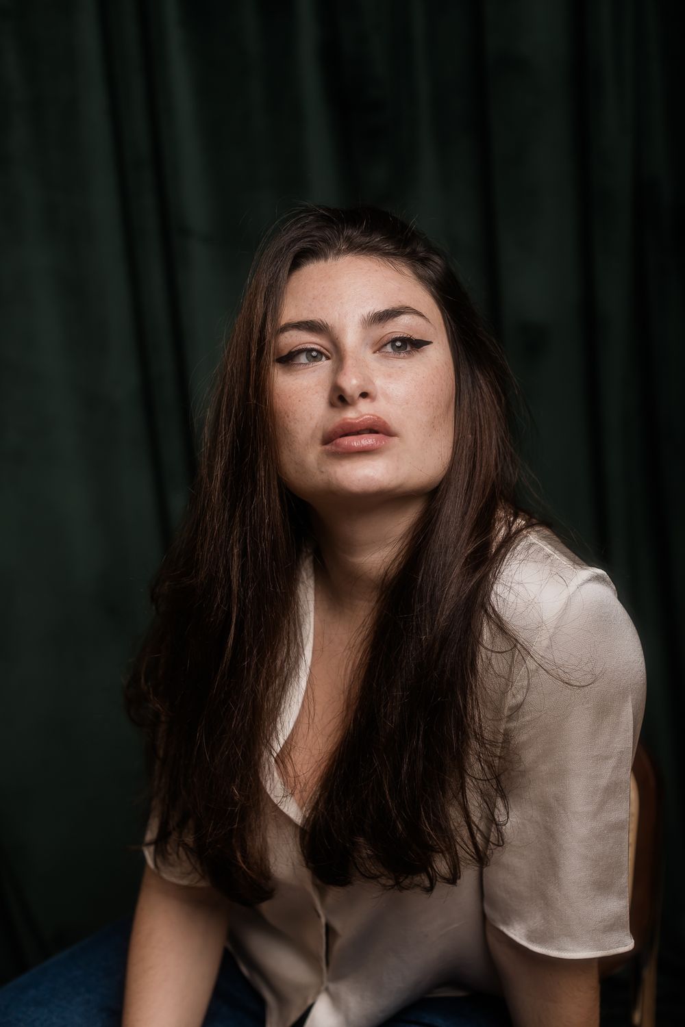 Moody portrait photograph of someone with long dark hair wearing a beige blouse against a dark green background.