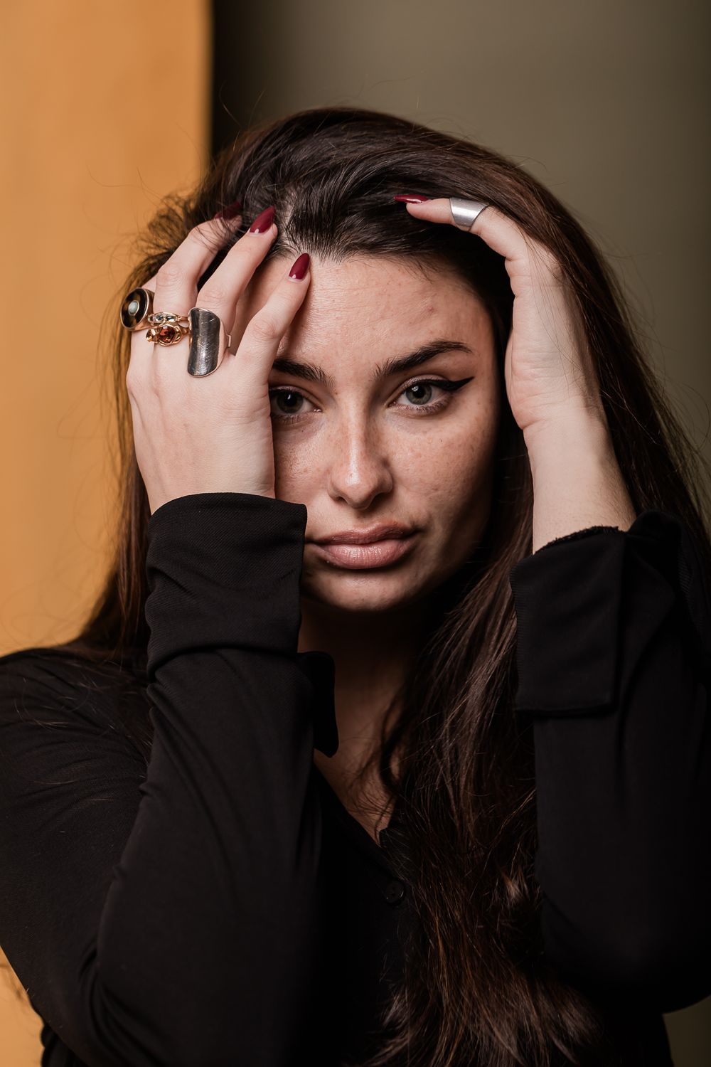 Portrait of person in black sweater with dark hair holding hands to head, wearing statement rings against orange background.