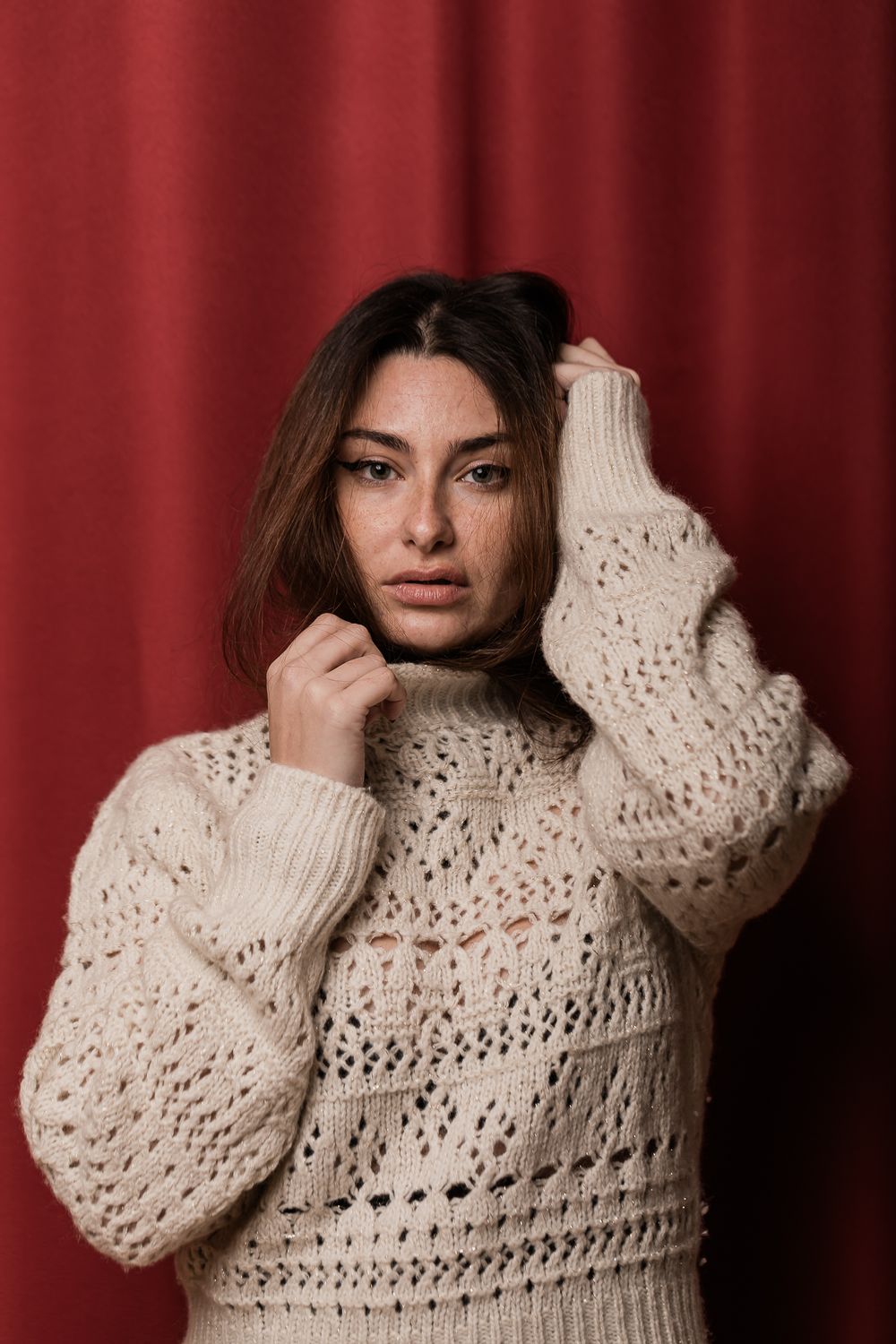 Model in cream knit sweater poses against red backdrop showing textured pattern detail.