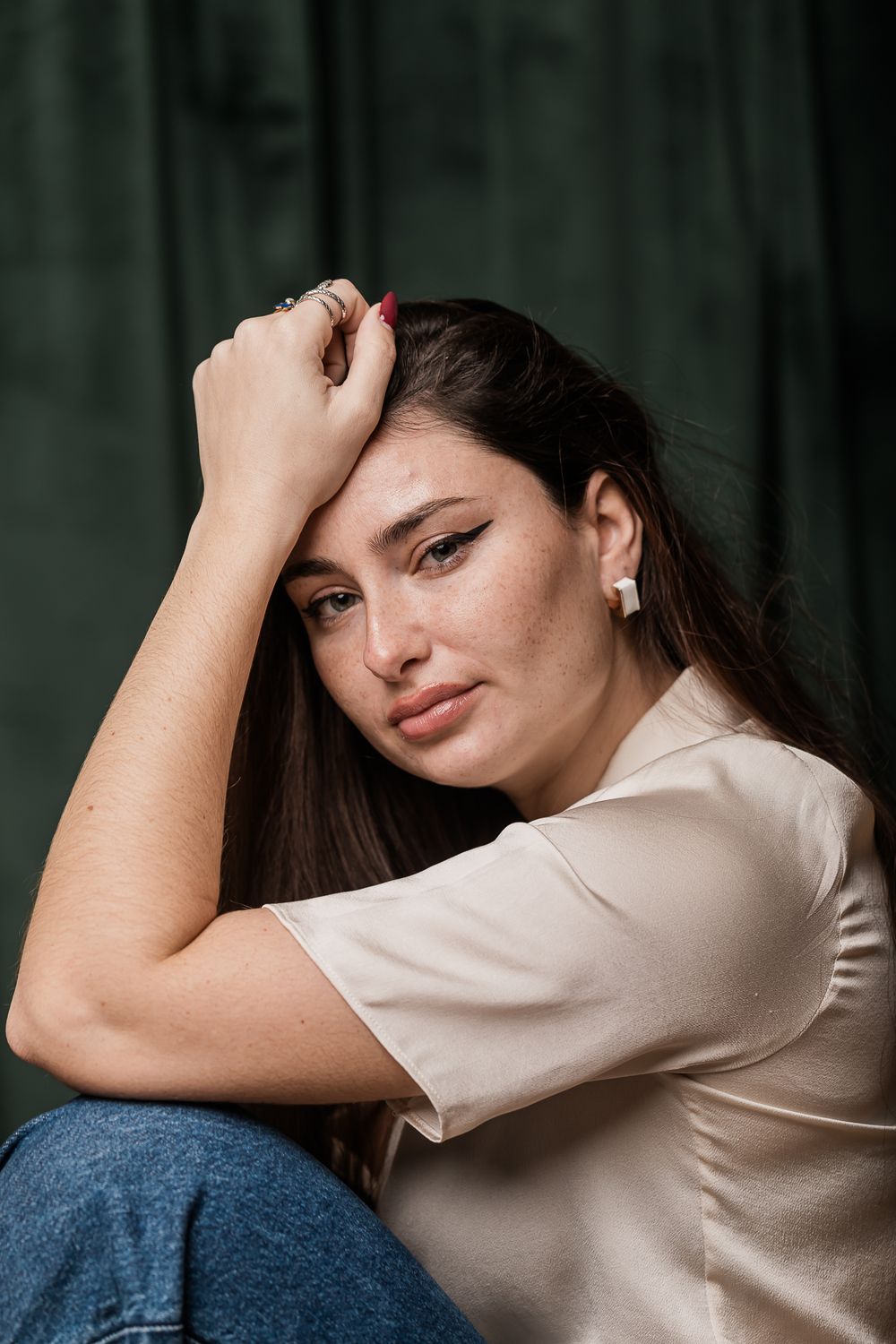 Artistic portrait of someone wearing a cream colored top against a dark green background in moody lighting.