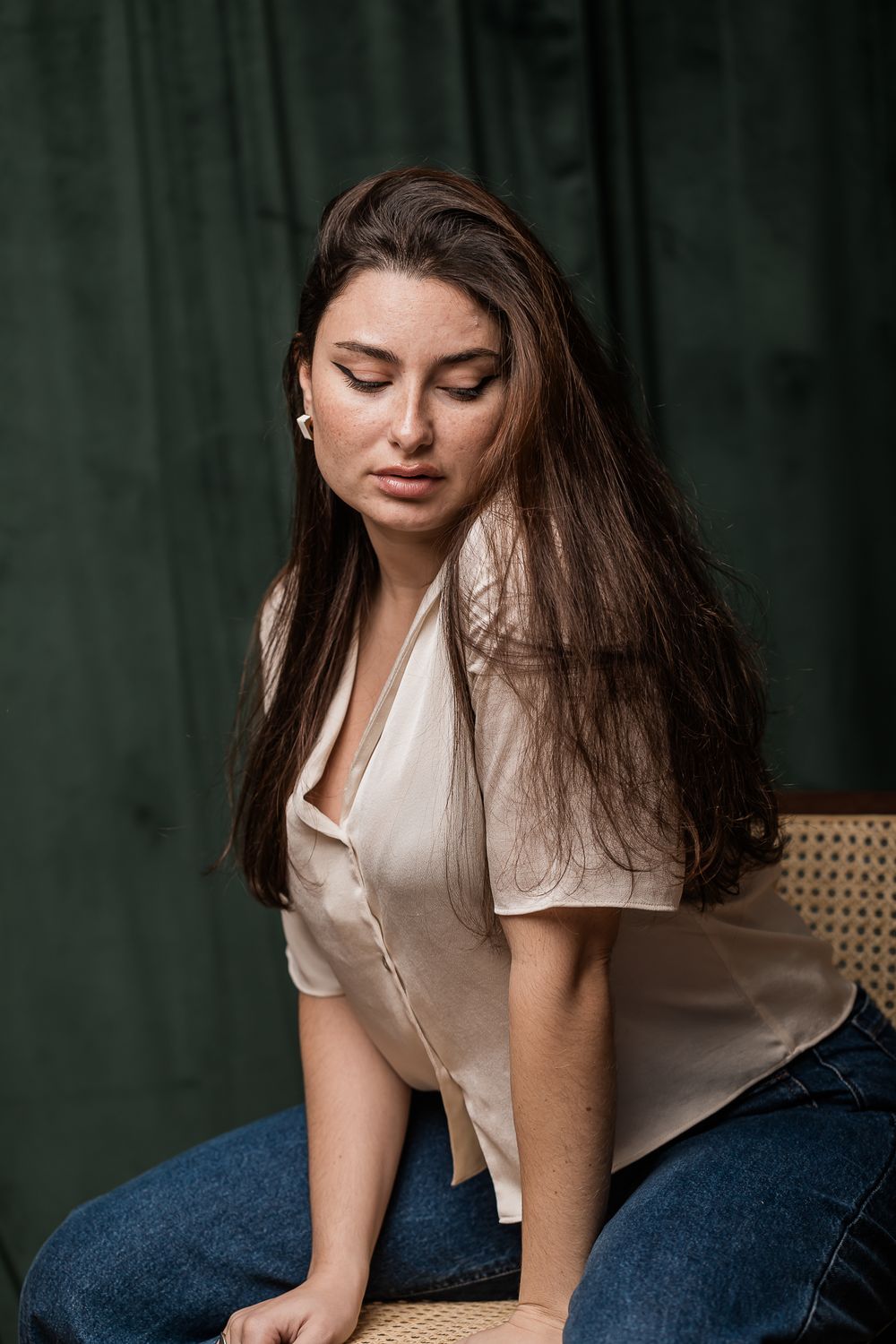 A person with long dark hair wearing a white blouse and jeans sits thoughtfully against a dark green background.