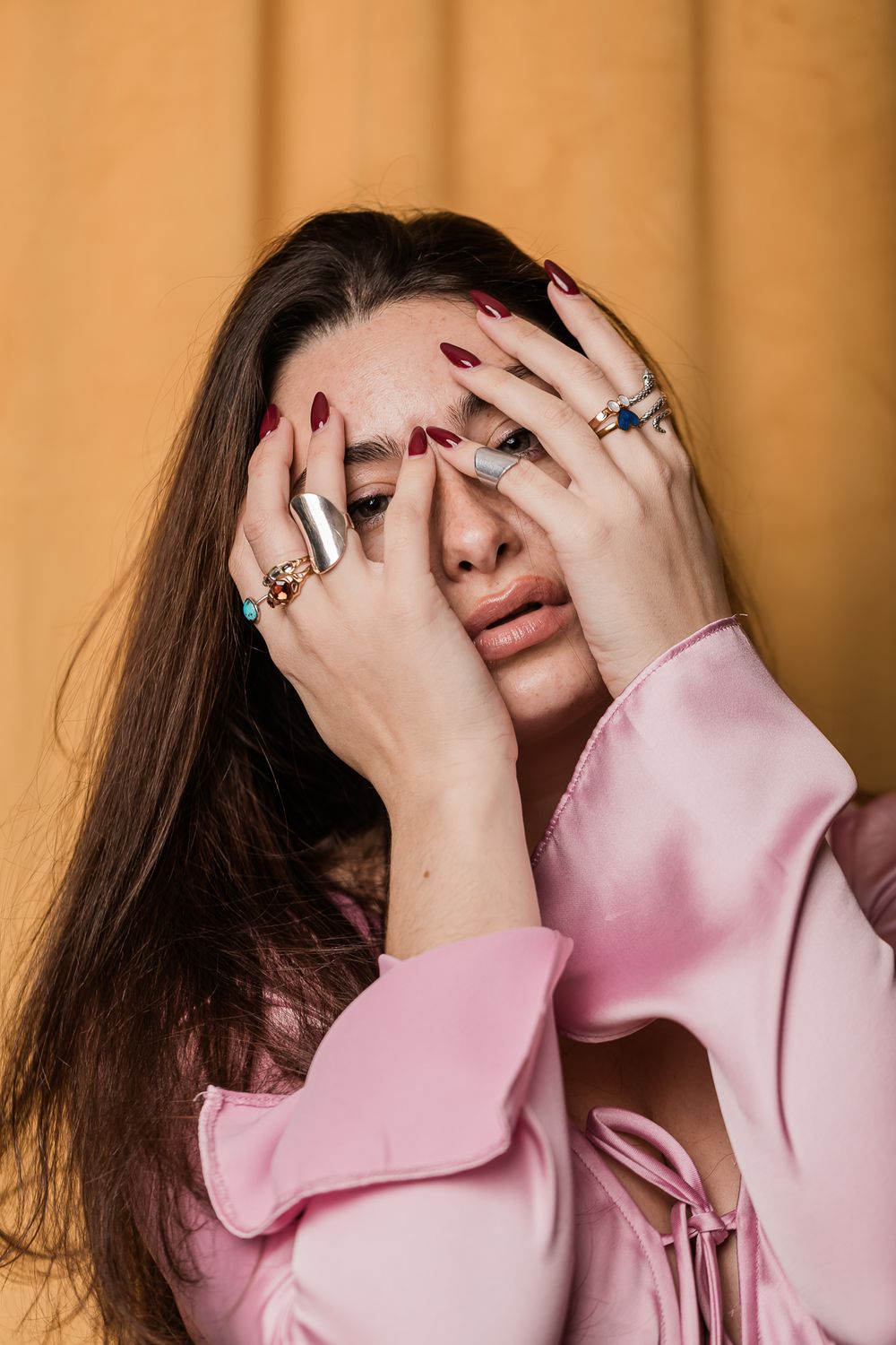 Person in pink sweater covering face with hands wearing multiple rings against beige background.