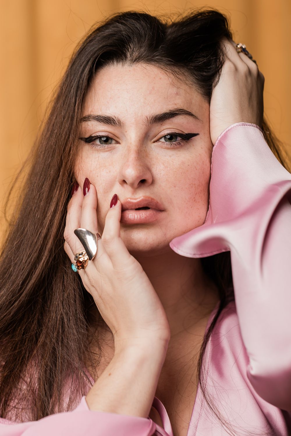 Fashion portrait featuring light pink blazer, statement ring and bold winged eyeliner against warm orange background.