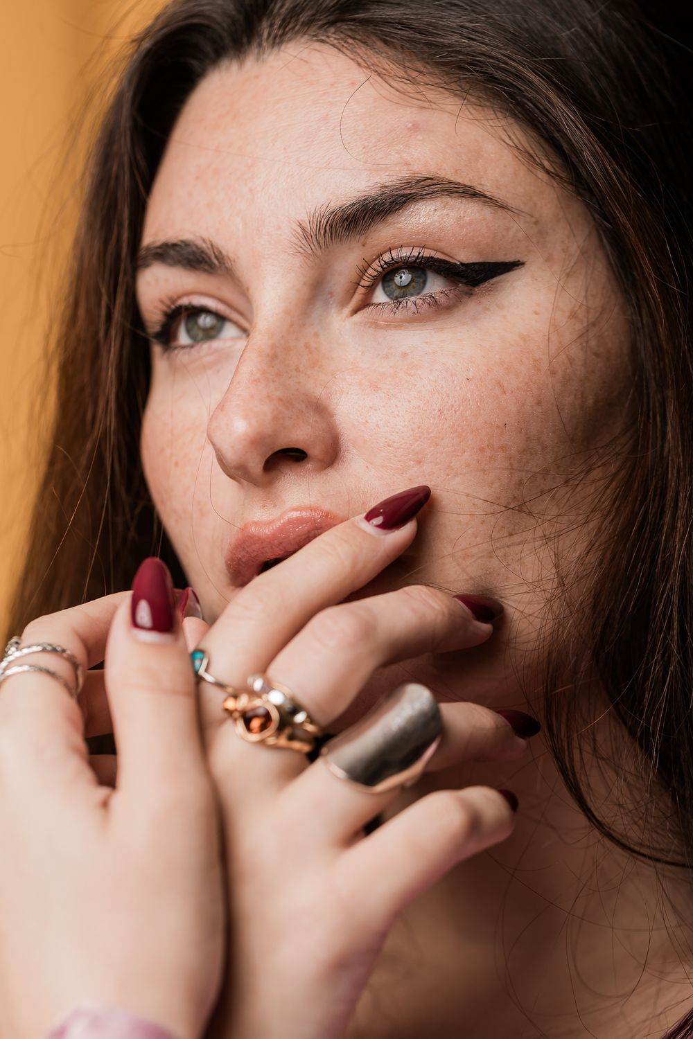 Close up beauty portrait featuring dramatic winged eyeliner, red lipstick, burgundy nails and multiple statement rings.