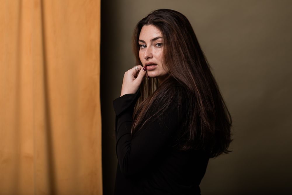 Woman with long dark hair poses against orange background wearing black sweater in moody portrait.