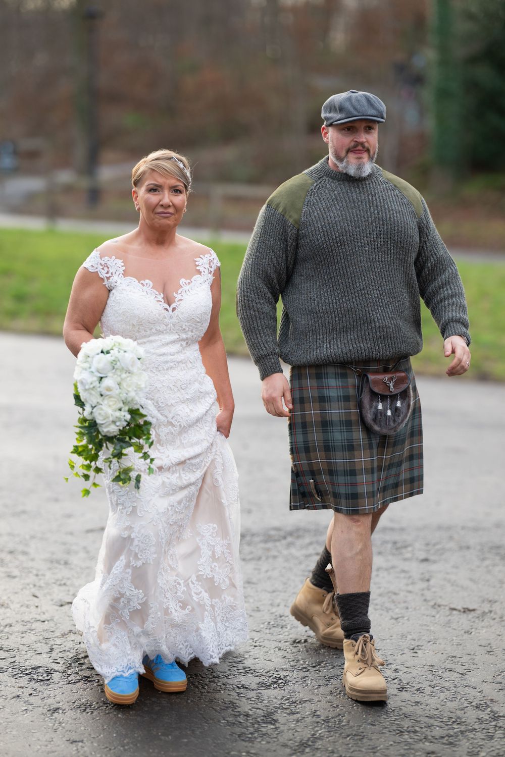 Bride in white lace dress and groom in Scottish kilt walking together outdoors.