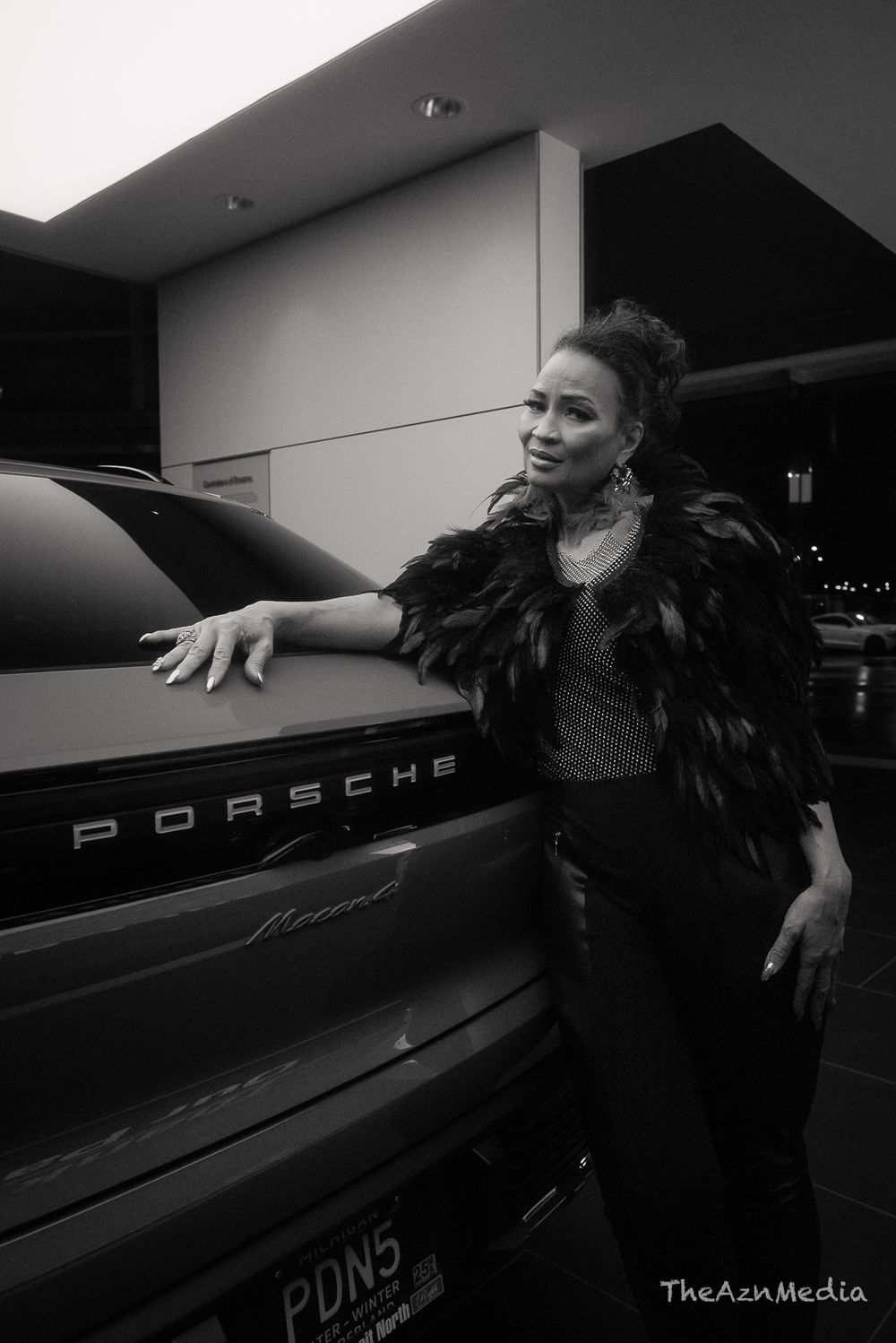 Stylish person in black feathered jacket poses against a luxury Porsche vehicle.