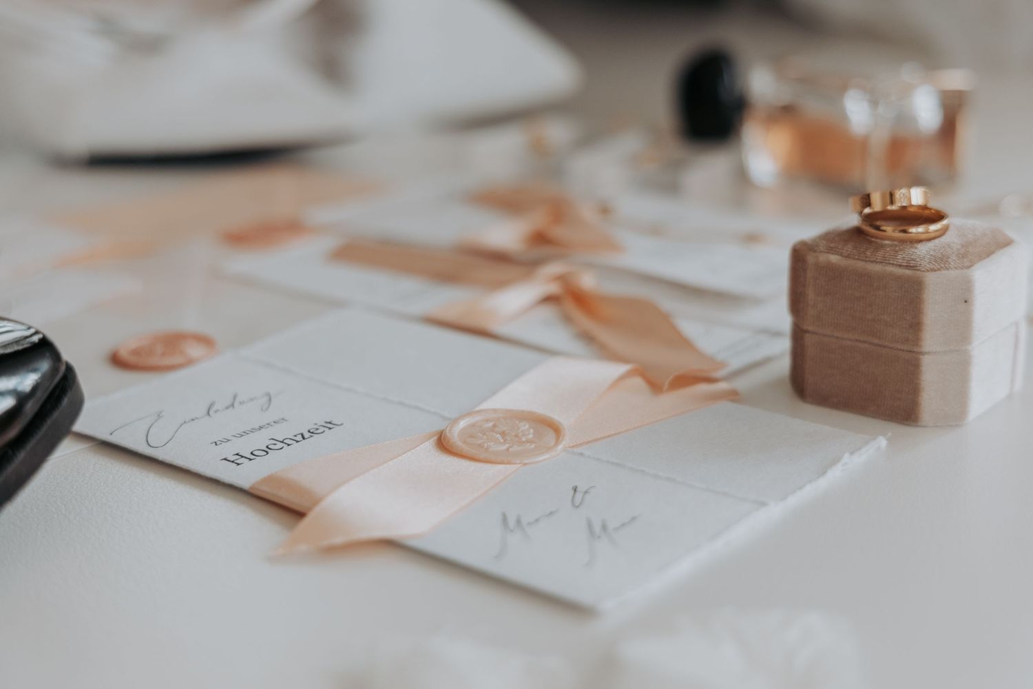 Elegant wedding invitations with wax seals and a small gift box in rose gold tones lay on white surface.