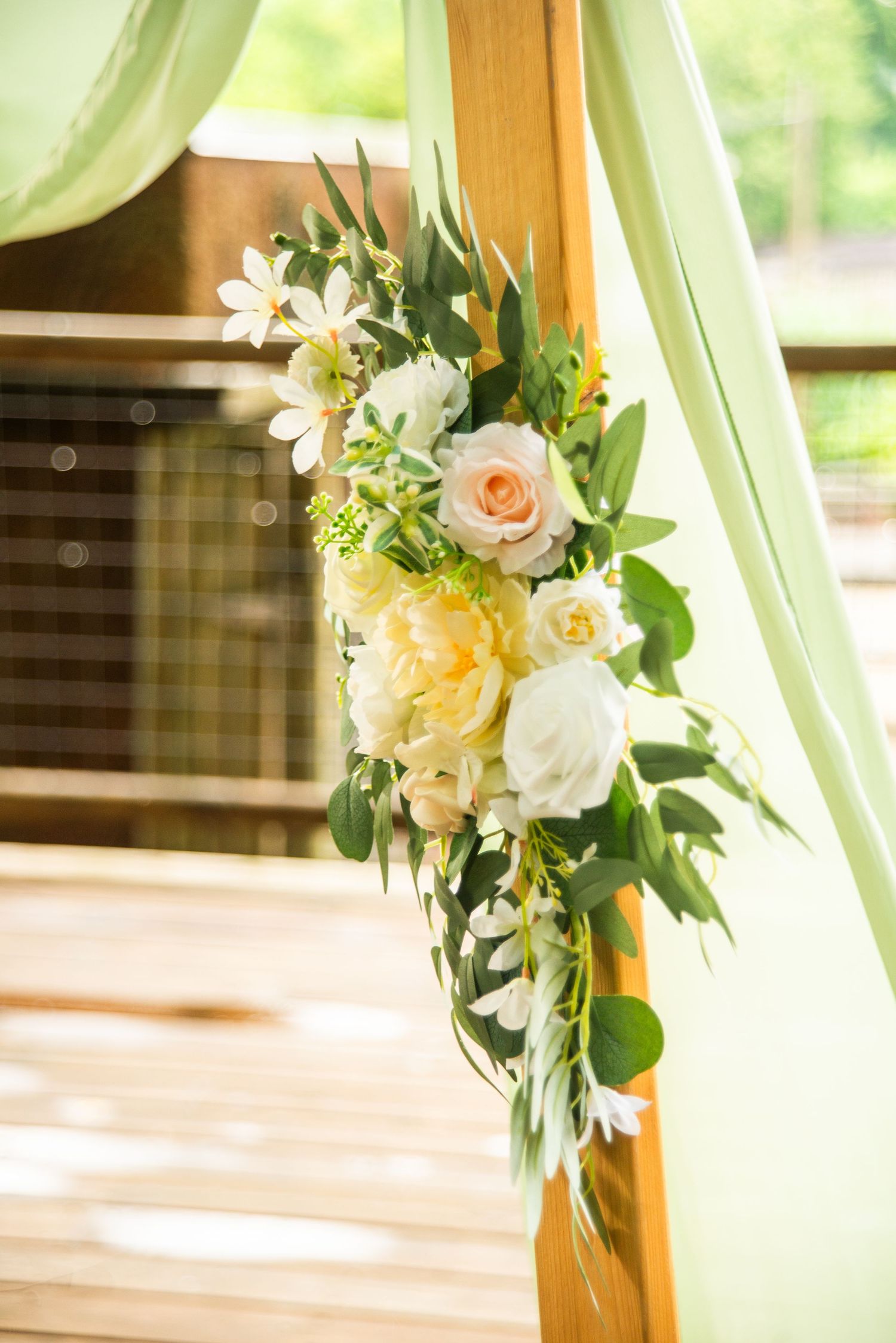 A cascading floral arrangement with peach roses, white lilies and greenery hangs elegantly on a wooden post with mint green fabric.