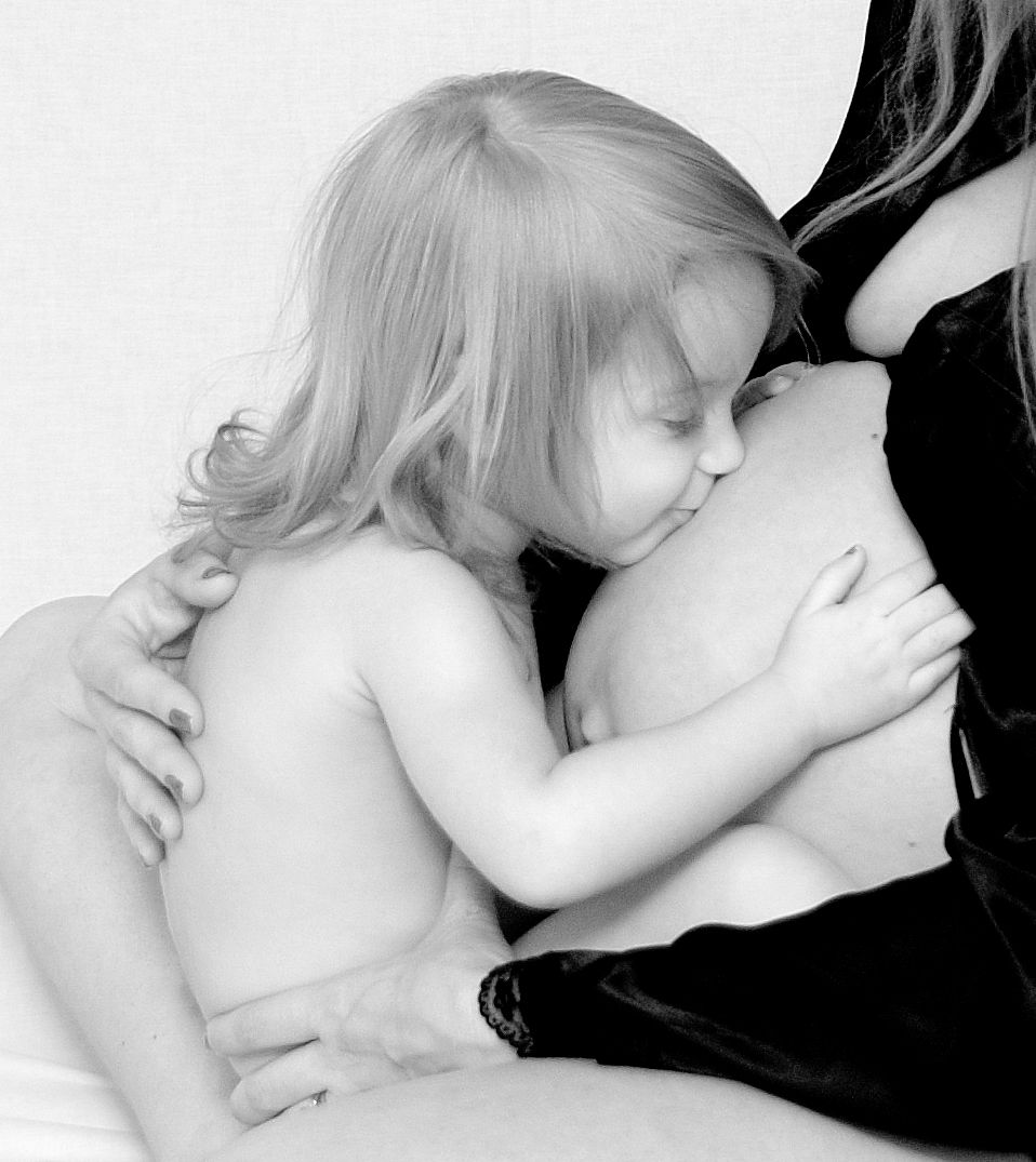 A black and white intimate photograph showing a small toddler resting against a pregnant belly in tender embrace.
