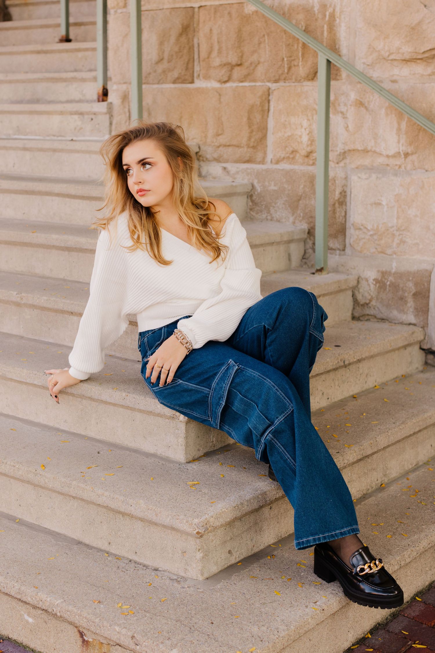 Casual fashion portrait on stone steps showing white sweater, wide-leg jeans and black loafers in warm sunlight.