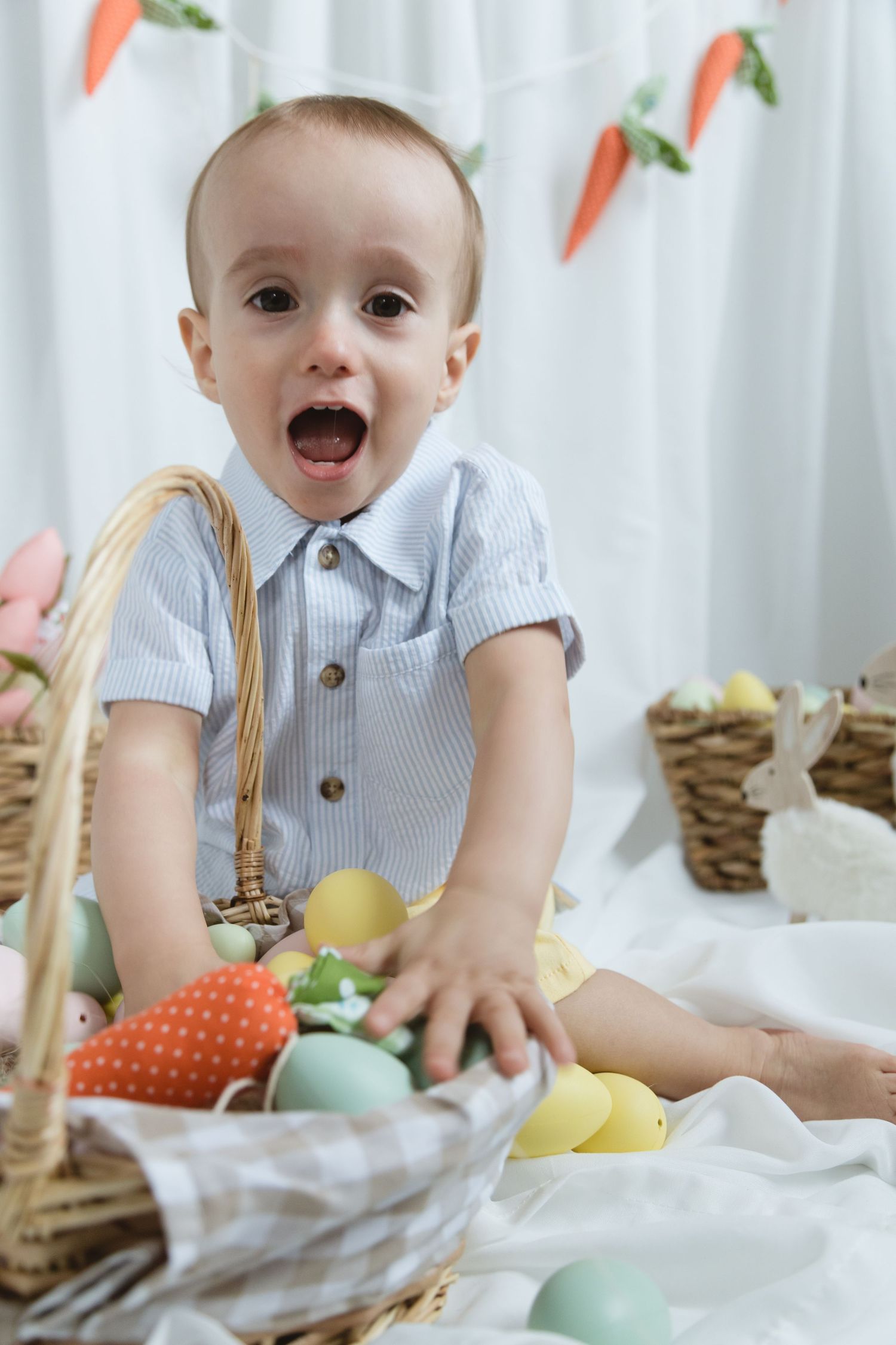 A baby wearing a blue striped outfit sits with Easter decorations and baskets surrounded by carrot decorations on white backdrop.
