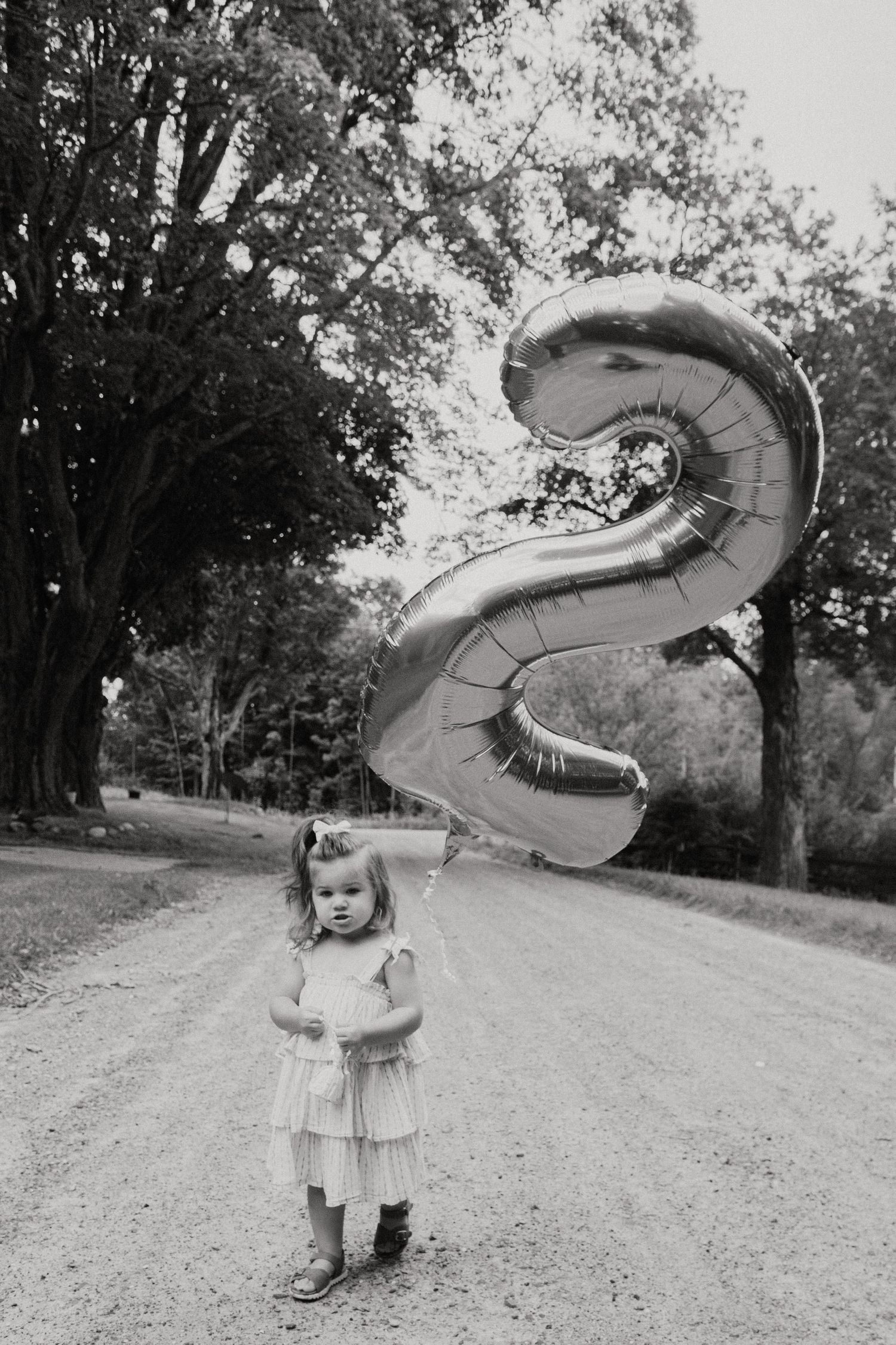 A child in a dress holds a large silver number 2 balloon on a tree-lined dirt road.
