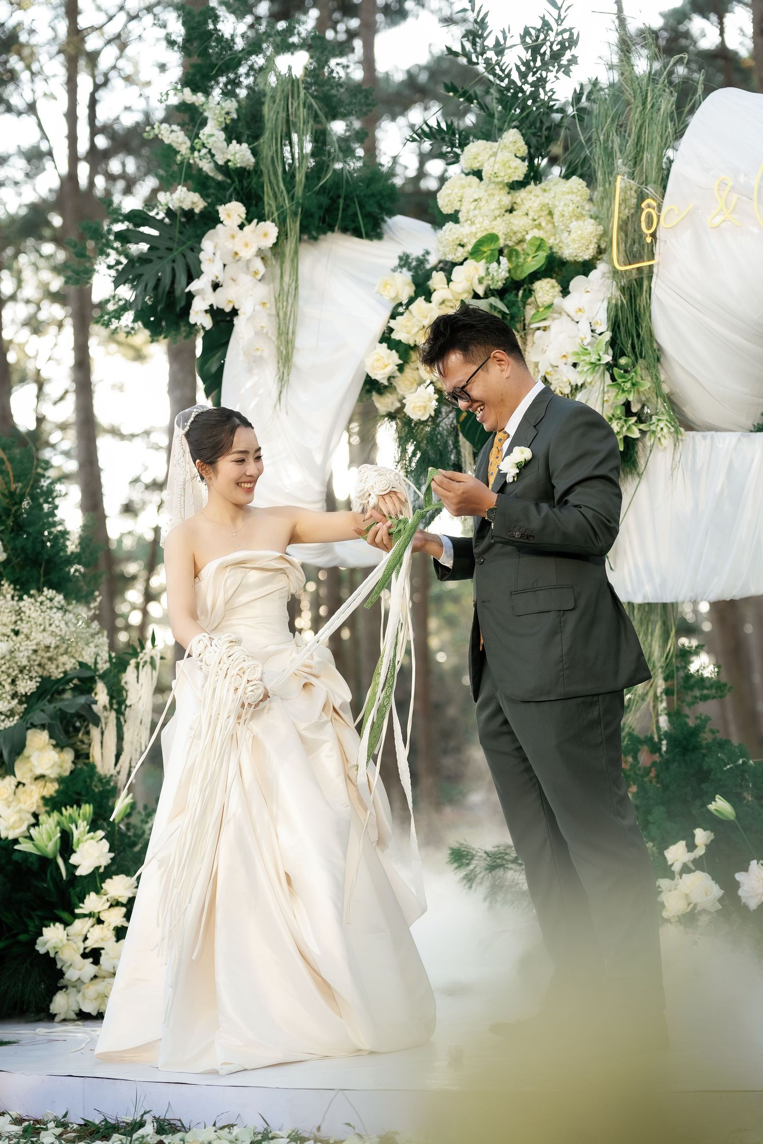 An elegant outdoor wedding ceremony features a floral arch with white and yellow blooms against a backdrop of pine trees.