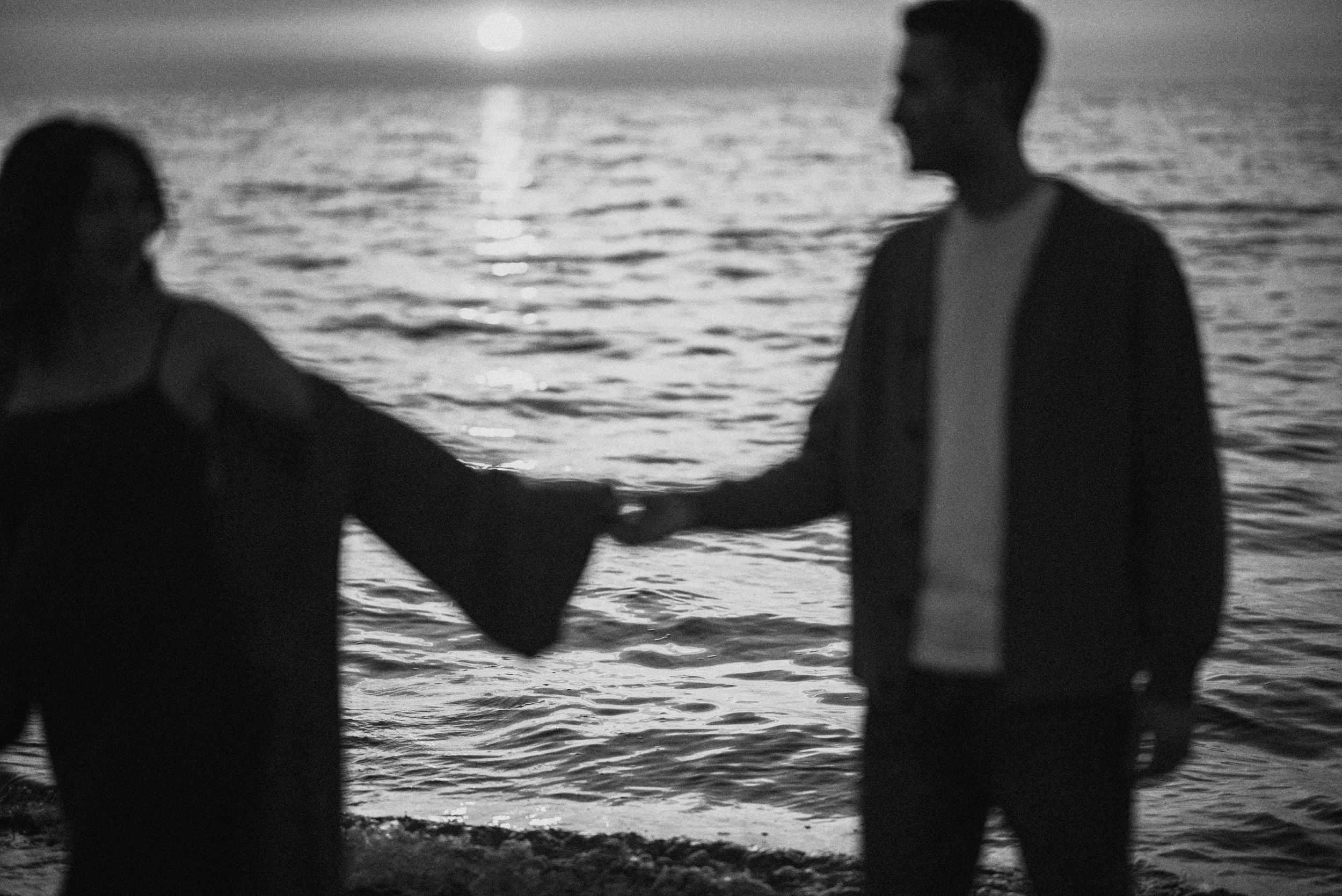 Black and white silhouette of two people holding hands by the ocean at sunset.
