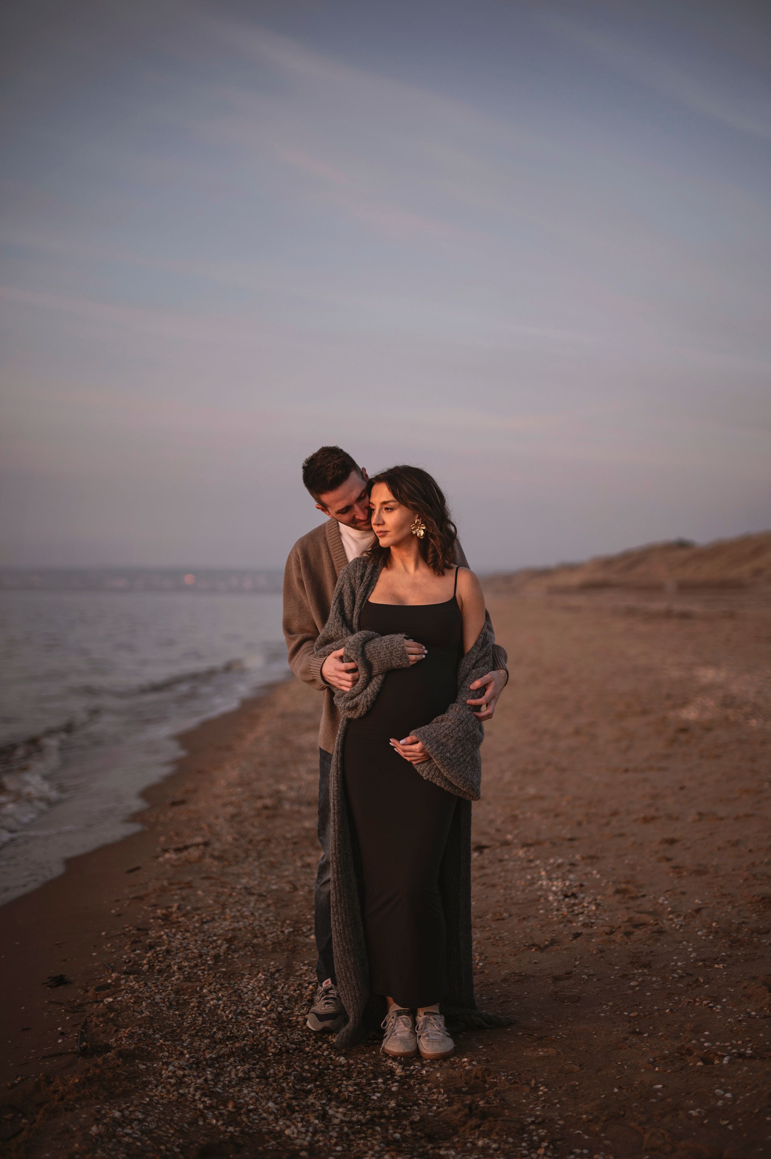 A couple embraces on a beach at dusk with soft lighting and a pregnant figure in a flowing black dress.